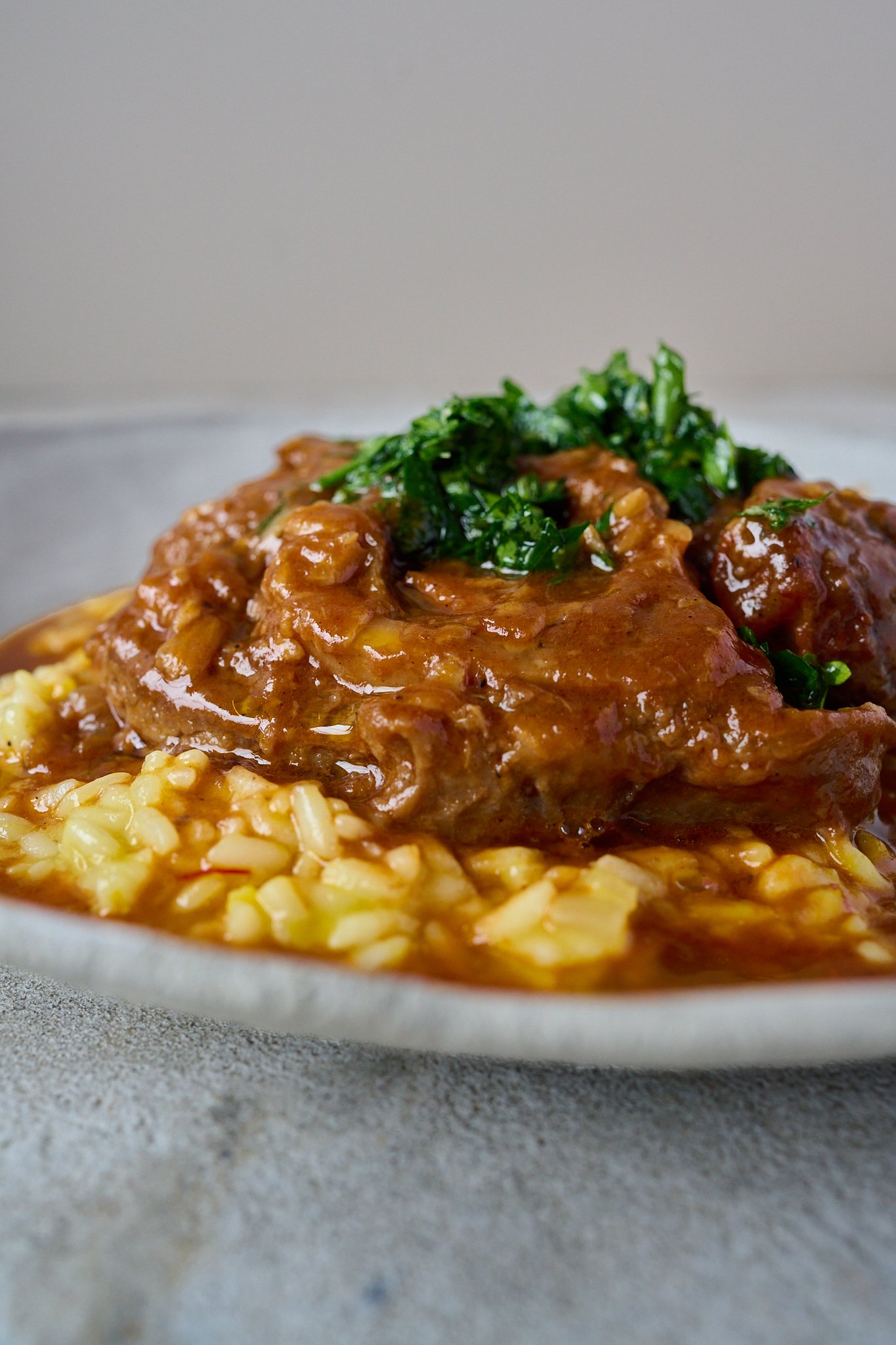 Osso buco sits on a bed of saffron risotto in a large white bowl with gremolata spooned on top. lots of sauce has pooled at the bottom. 