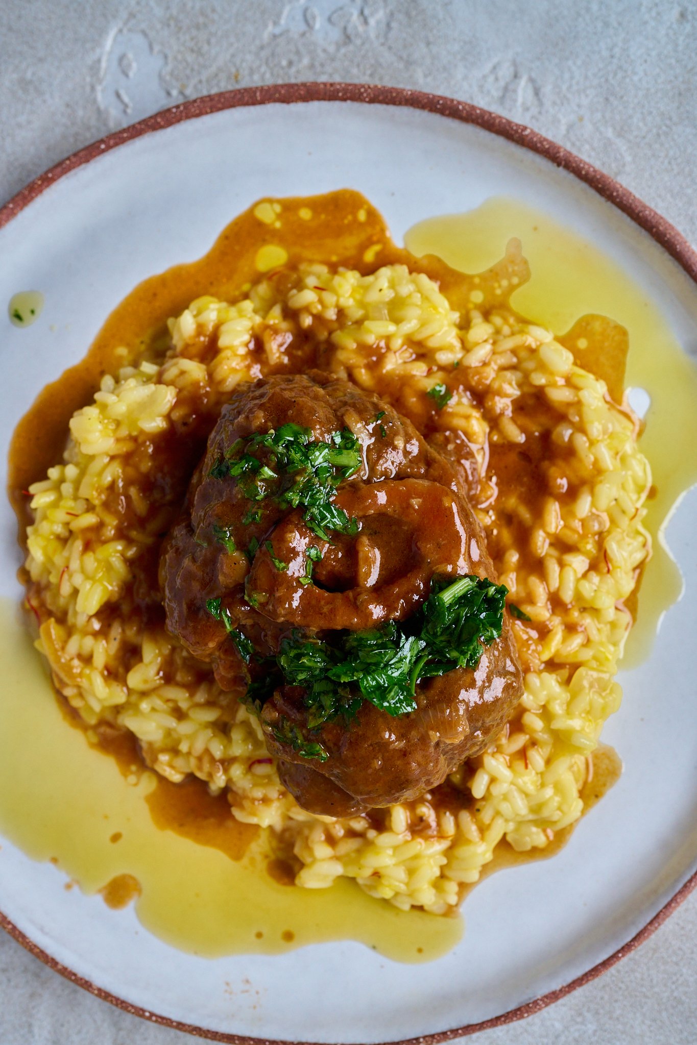 Osso buco sits on a bed of saffron risotto in a large white bowl with gremolata spooned on top. lots of sauce has pooled at the bottom. 