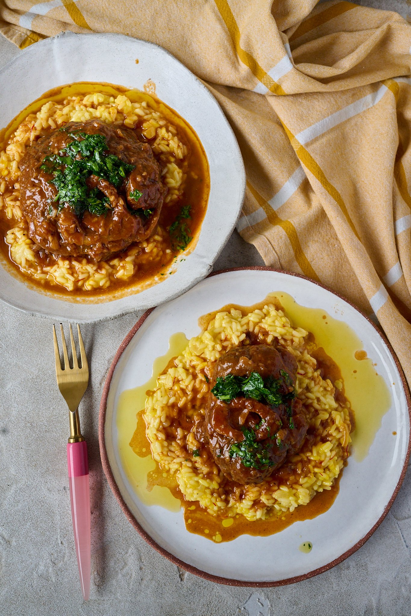 You can see two white bowls with Osso buco siting on a bed of saffron risotto in a large white bowl with gremolata spooned on top. lots of sauce has pooled at the bottom on both plates. there is a pink fork and orange and white tea towel. 