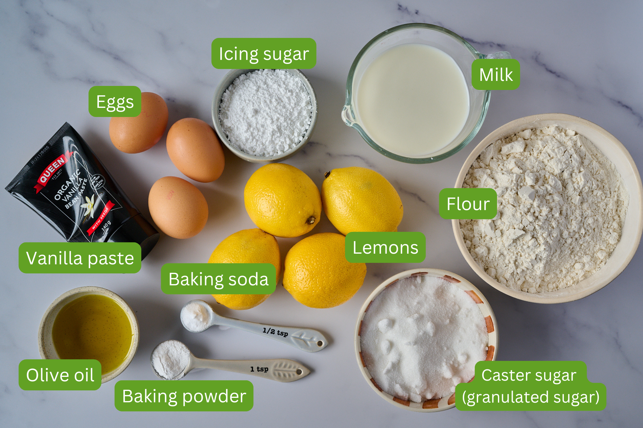 List of ingredients for lemon olive oil cake are laid out onto marble backdrop. Ingredients include eggs, vanilla paste, olive oil, baking powder, baking soda, lemons, caster sugar, flour, milk and icing sugar