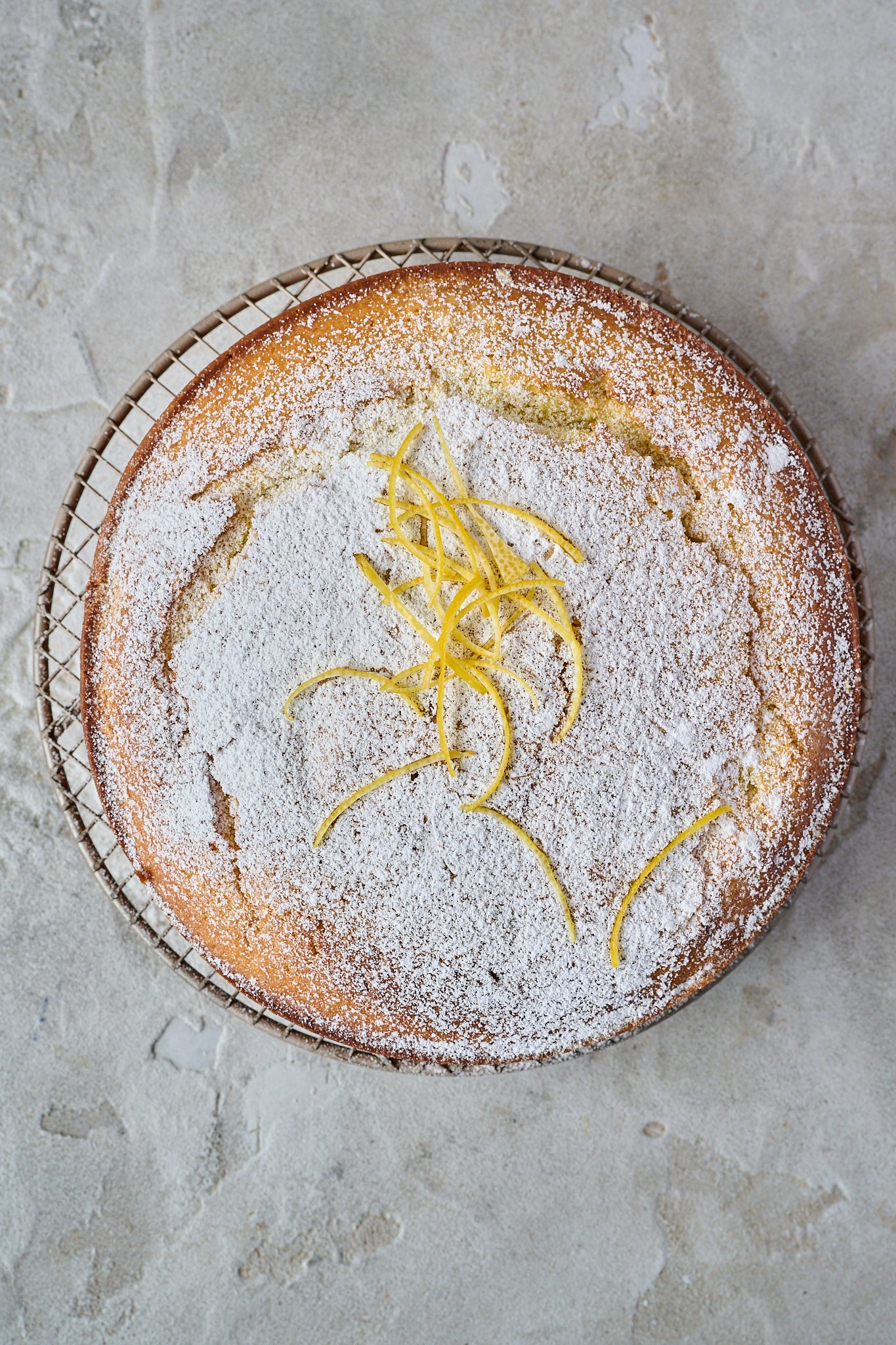 Lemon olive oil cake has been transferred to a round cooling rack. It's dusted with icing sugar and has lemon zest sitting on top. 