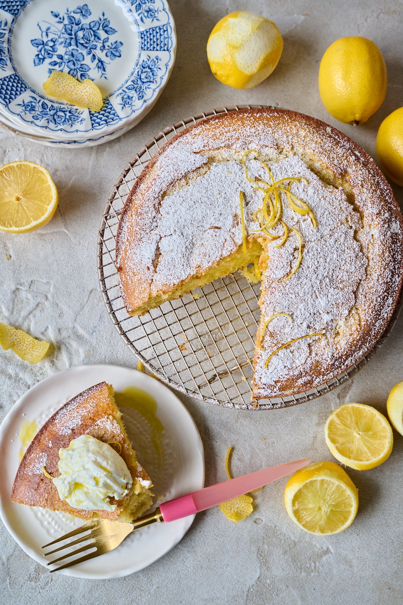 Sliced lemon olive oil cake sits on round cooling rack. Once slice is taken out of it and is sitting on a white plate in front of it. Cut up lemons are sitting around the cake and there is a pink fork. 