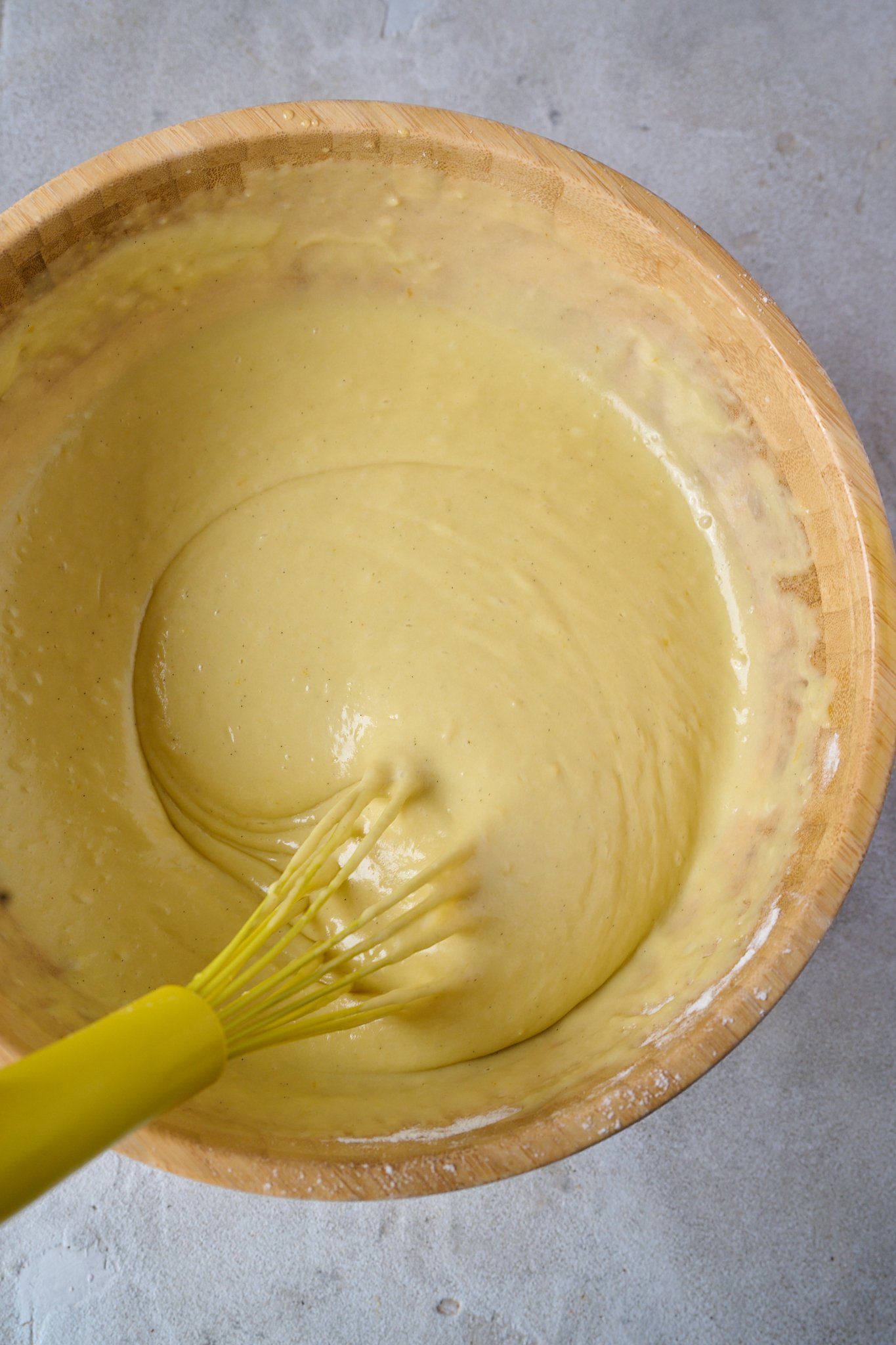 Batter for lemon olive oil cake sits in wooden bowl with yellow whisk sitting out the side