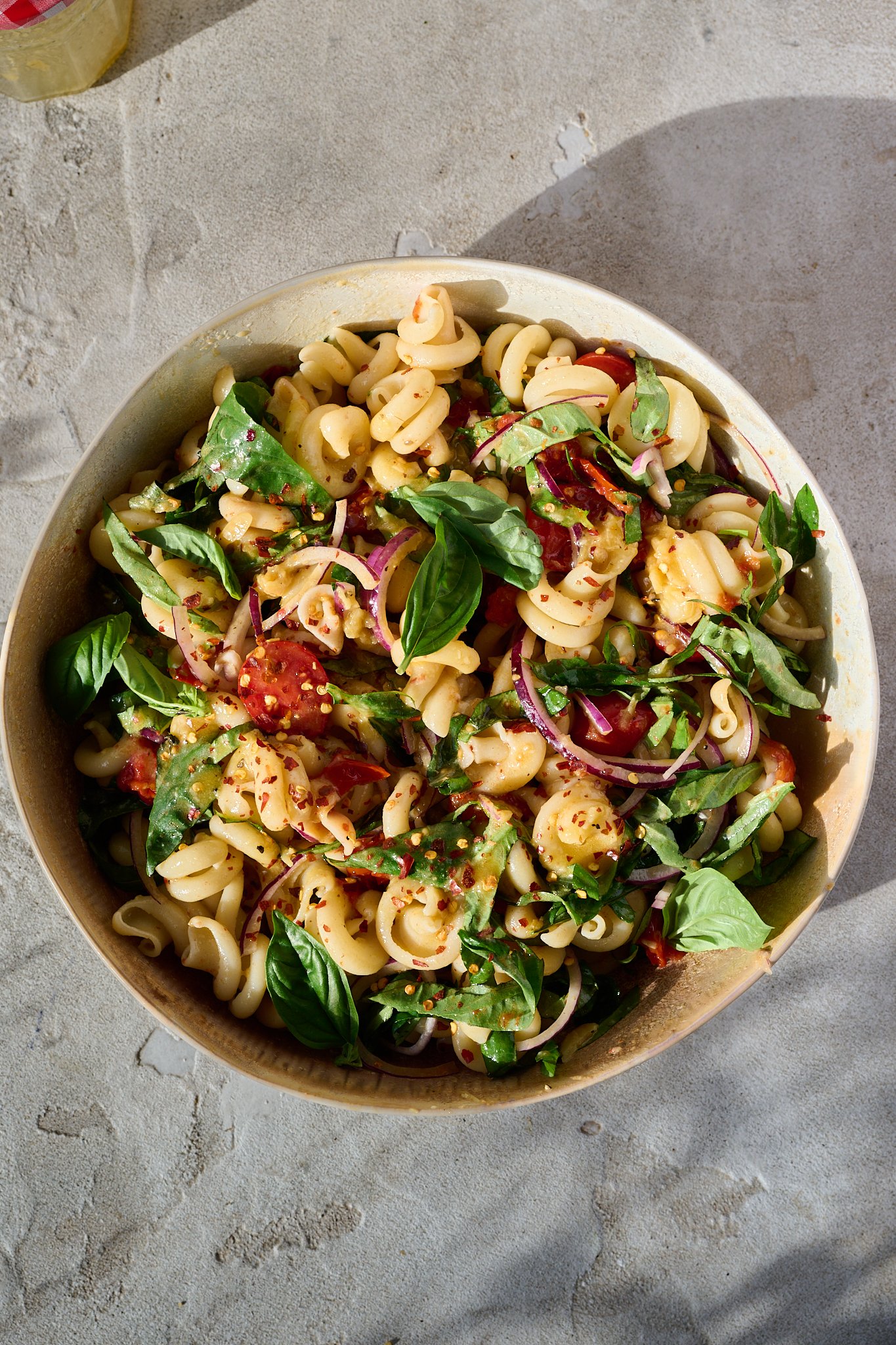 Mediterranean Tomato Confit Pasta Salad sits in large bowl in the sun. There are fresh basil leaves and chilli flakes sprinkled on top.