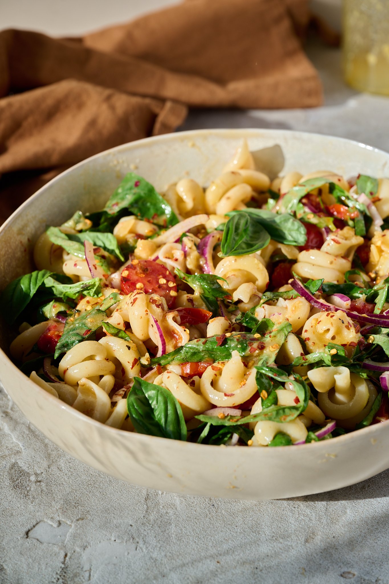 Mediterranean Tomato Confit Pasta Salad sits in large bowl in the sun. There are fresh basil leaves and chilli flakes sprinkled on top.