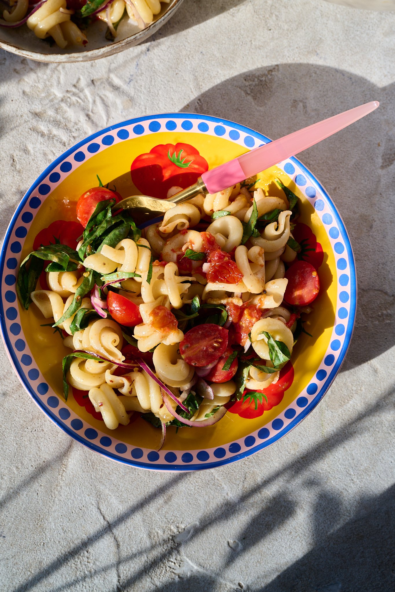 Mediterranean Tomato Confit Pasta Salad sits in large bowl in the sun. A pink fork sits out the side of the bowl. The bowl is yellow with a pink and blue rim. There are fresh basil leaves and chilli flakes sprinkled on top.