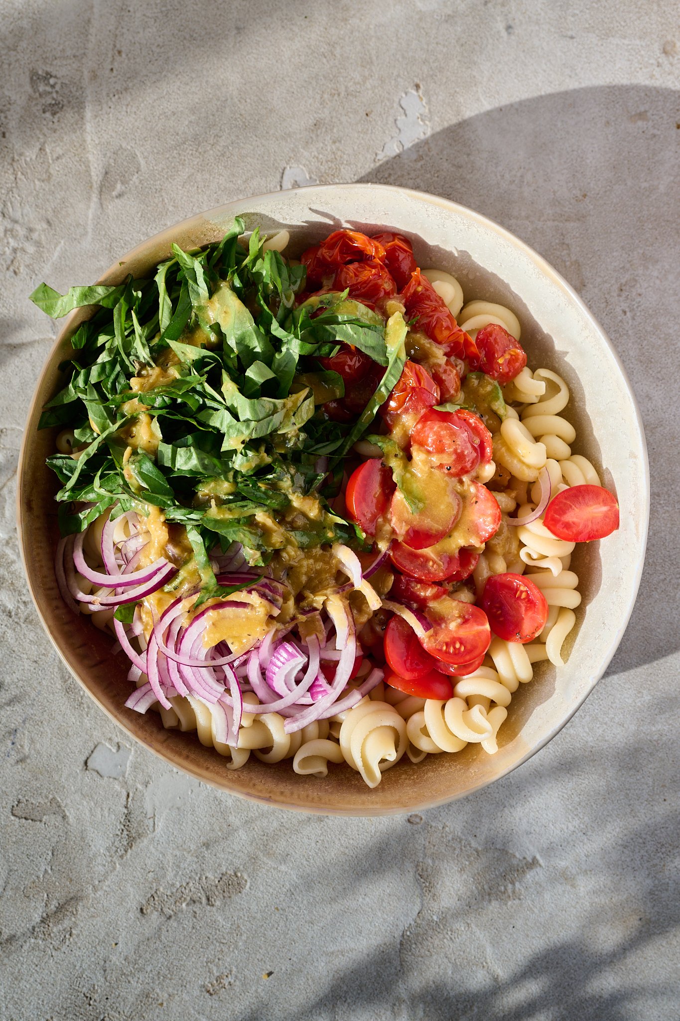 Mediterranean Tomato Confit Pasta Salad sits in large bowl in the sun. There are fresh basil leaves and chilli flakes sprinkled on top.