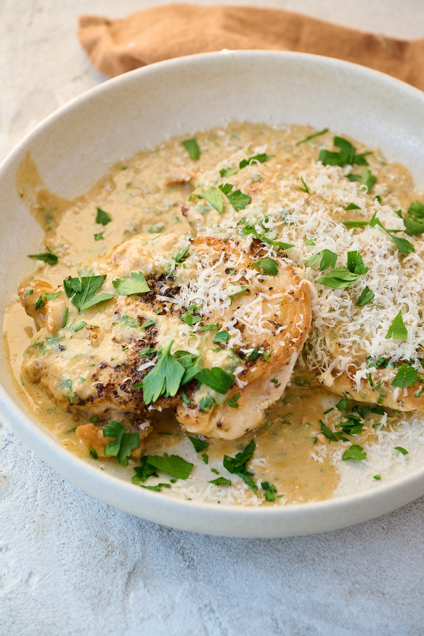 Creamy garlic parmesan chicken is served in white plate with fresh parsley and cheese sprinkled on top. 