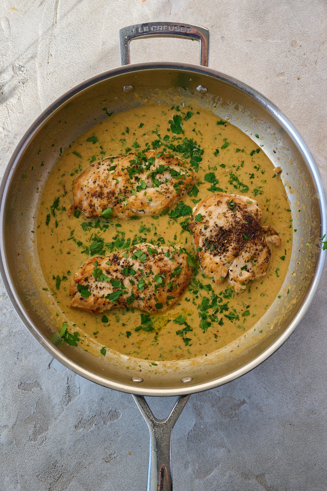 Creamy Garlic Parmesan Chicken sits in stainless steel pan with sauce all around it. Fresh parsley is sprinkled on top. 