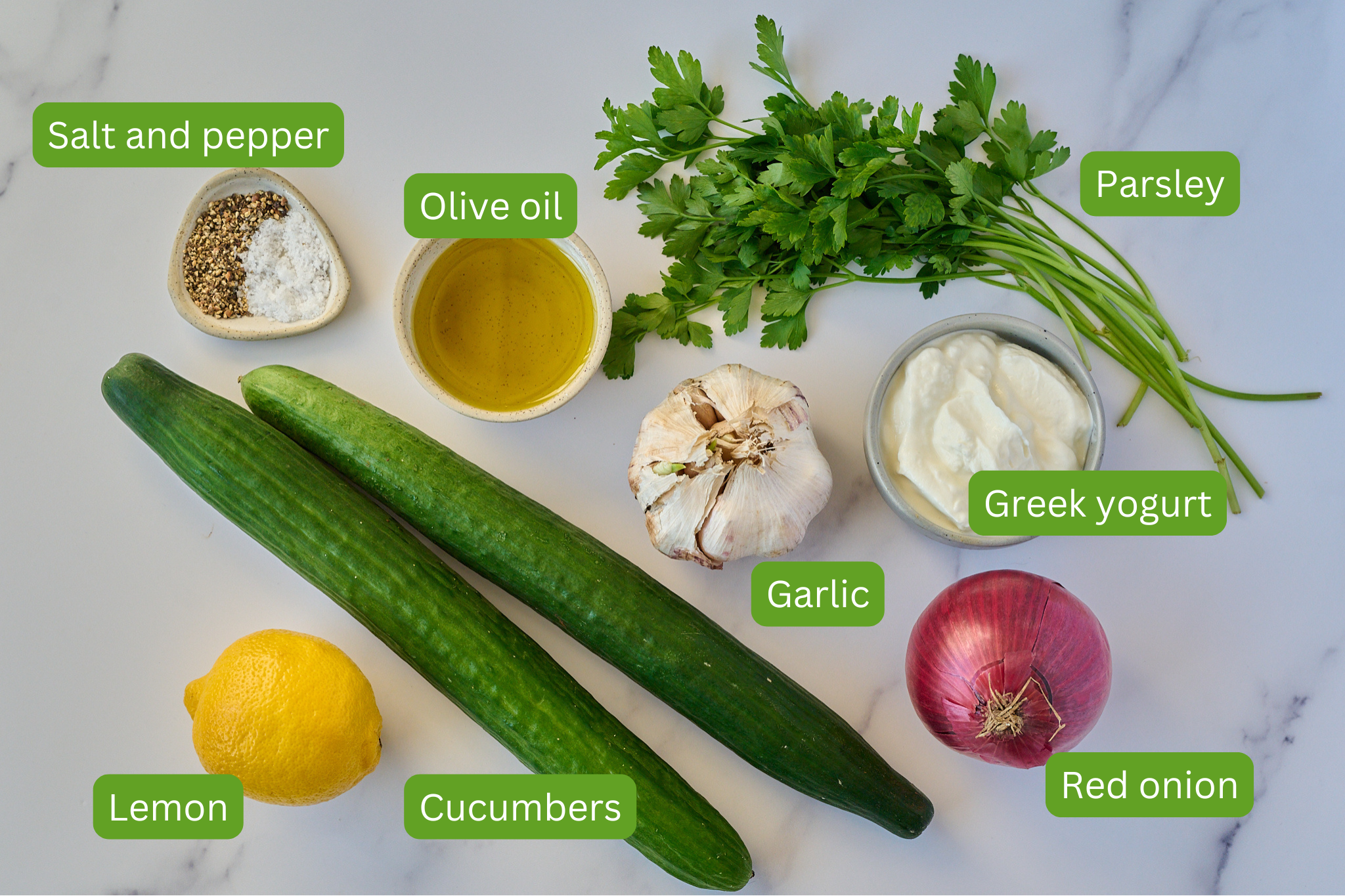 Ingredients for creamy cucumber salad are laid out on marble backdrop. You can see cucumbers, lemon, garlic, red onion, greek yogurt, parsley, olive oil and salt and pepper. 
