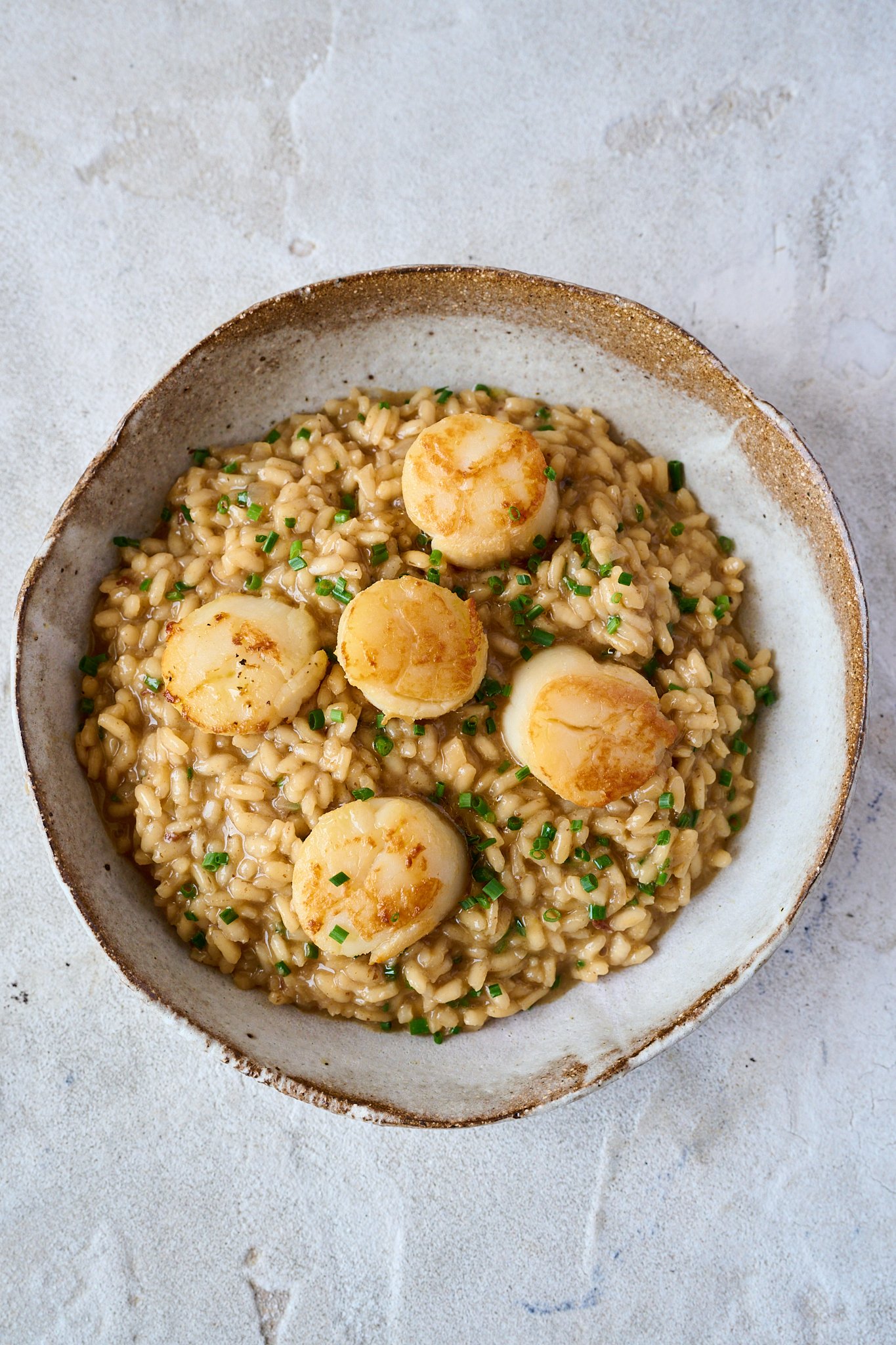 Brown bowl is filled with scallop risotto. Scallops are sitting on top of risotto and finished with a sprinkling of chives.