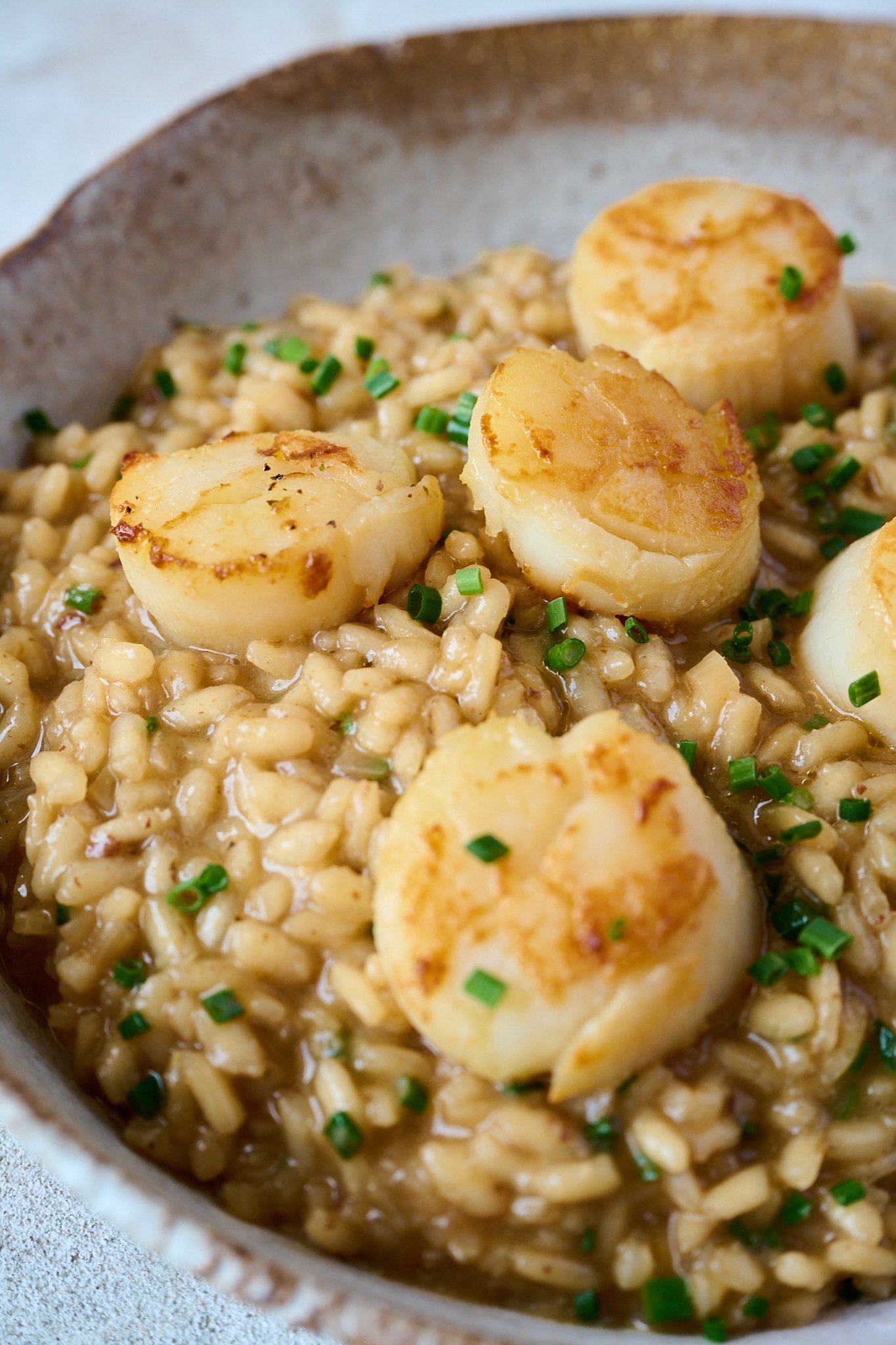 Brown bowl is filled with scallop risotto. Scallops are sitting on top of risotto and finished with a sprinkling of chives. 