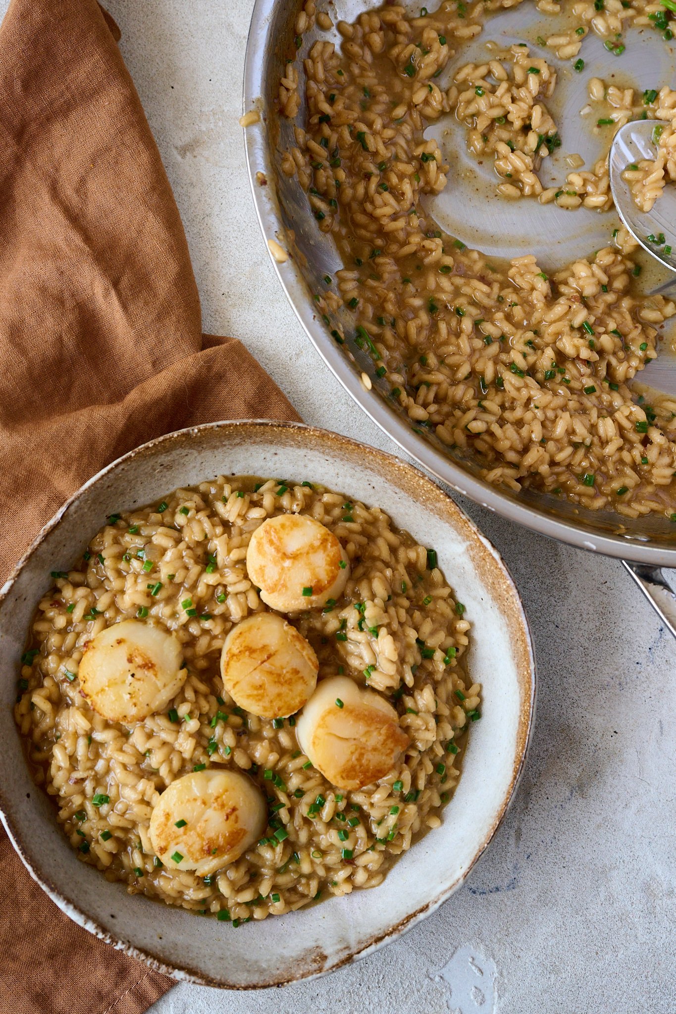Brown bowl is filled with scallop risotto. Scallops are sitting on top of risotto and finished with a sprinkling of chives. You can see a brown tea towel to the left of the bowl and a pan with risotto to the right. 