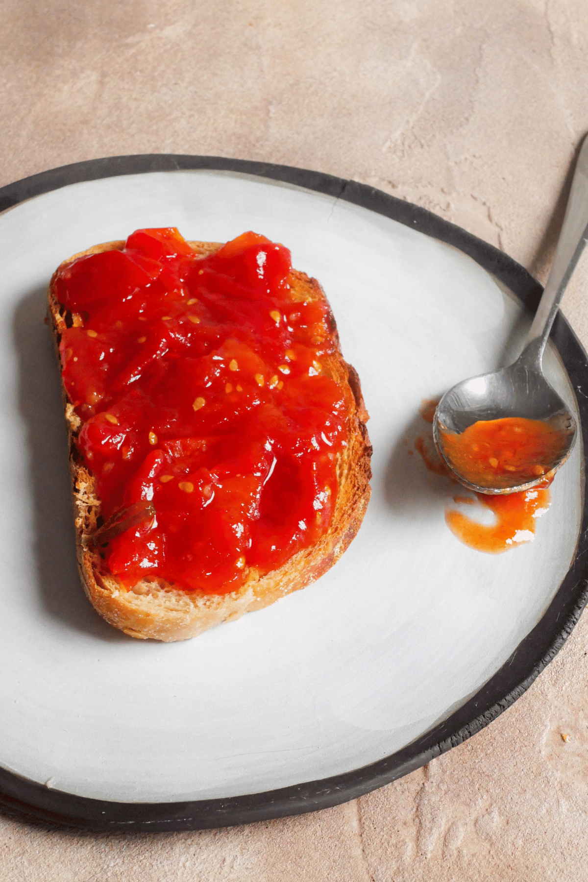 piece of toast lays on a plate with tomato jam spread all over it. Spoon with tomato jam is sticking out the side of the plate.