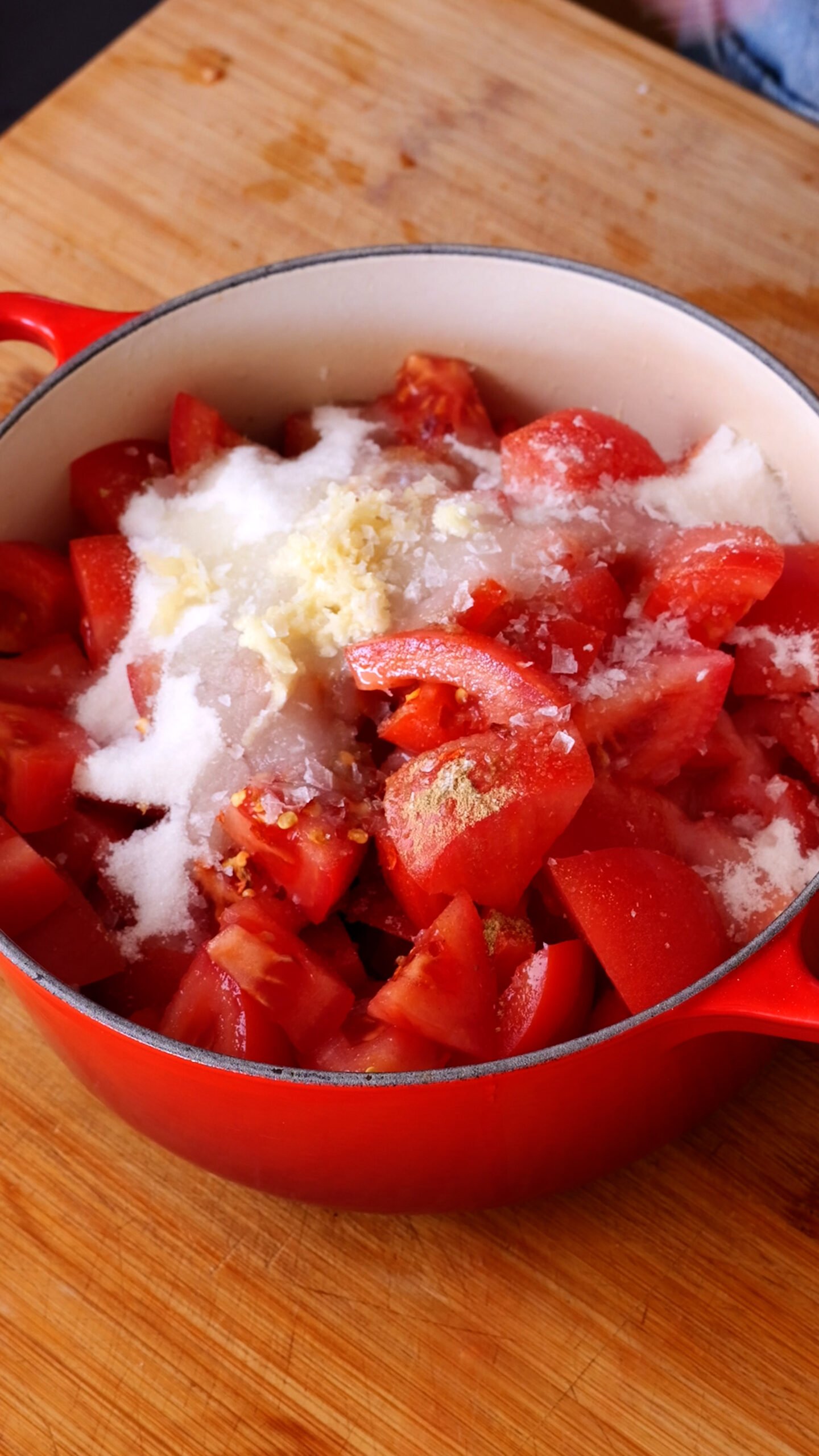 Red pot is filled with all ingredients to make tomato jam. You can see sugar, garlic, cumin, chilli flakes and tomatoes