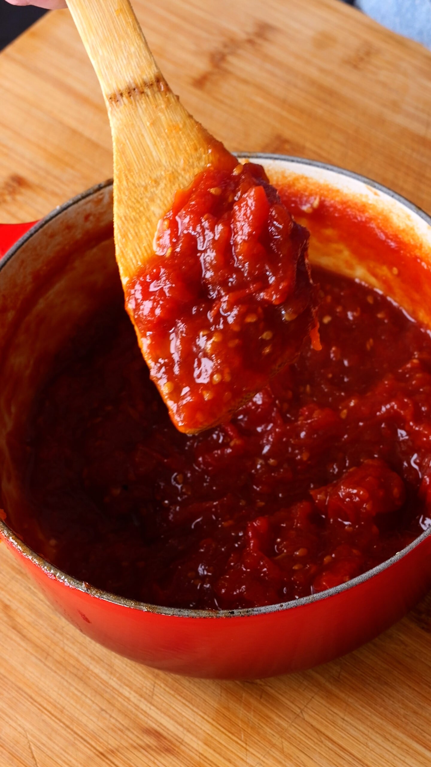 red pot is filled with cooked tomato jam and there is a wooden spoon with tomato jam all on it.