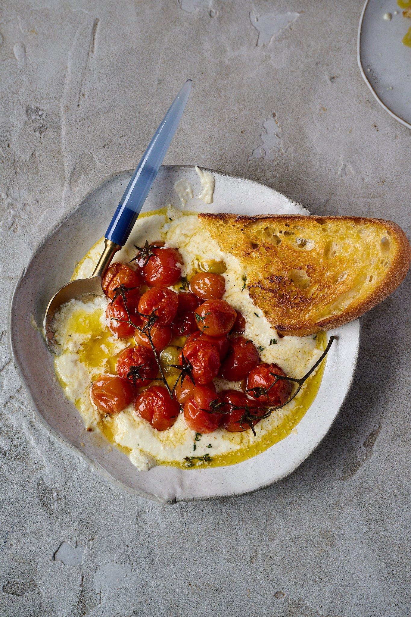 Large white plate is shown with whipped goat cheese and roasted tomatoes sitting on top. You can see a drizzle of honey and fresh thyme sprinkled all over. There is a blue spoon sticking out of the bowl on the left side and toasted sourdough on the right. 