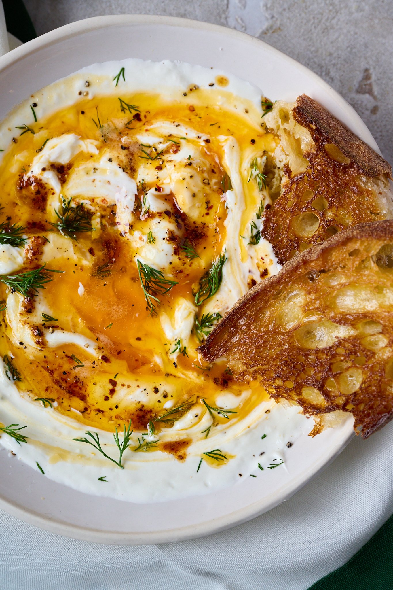 Turkish eggs are shown in white bowl. Two poached eggs have been broken open and sit on top of yoghurt with red butter sauce drizzled all over.  Two pieces of sourdough stick out the side of the bowl. 