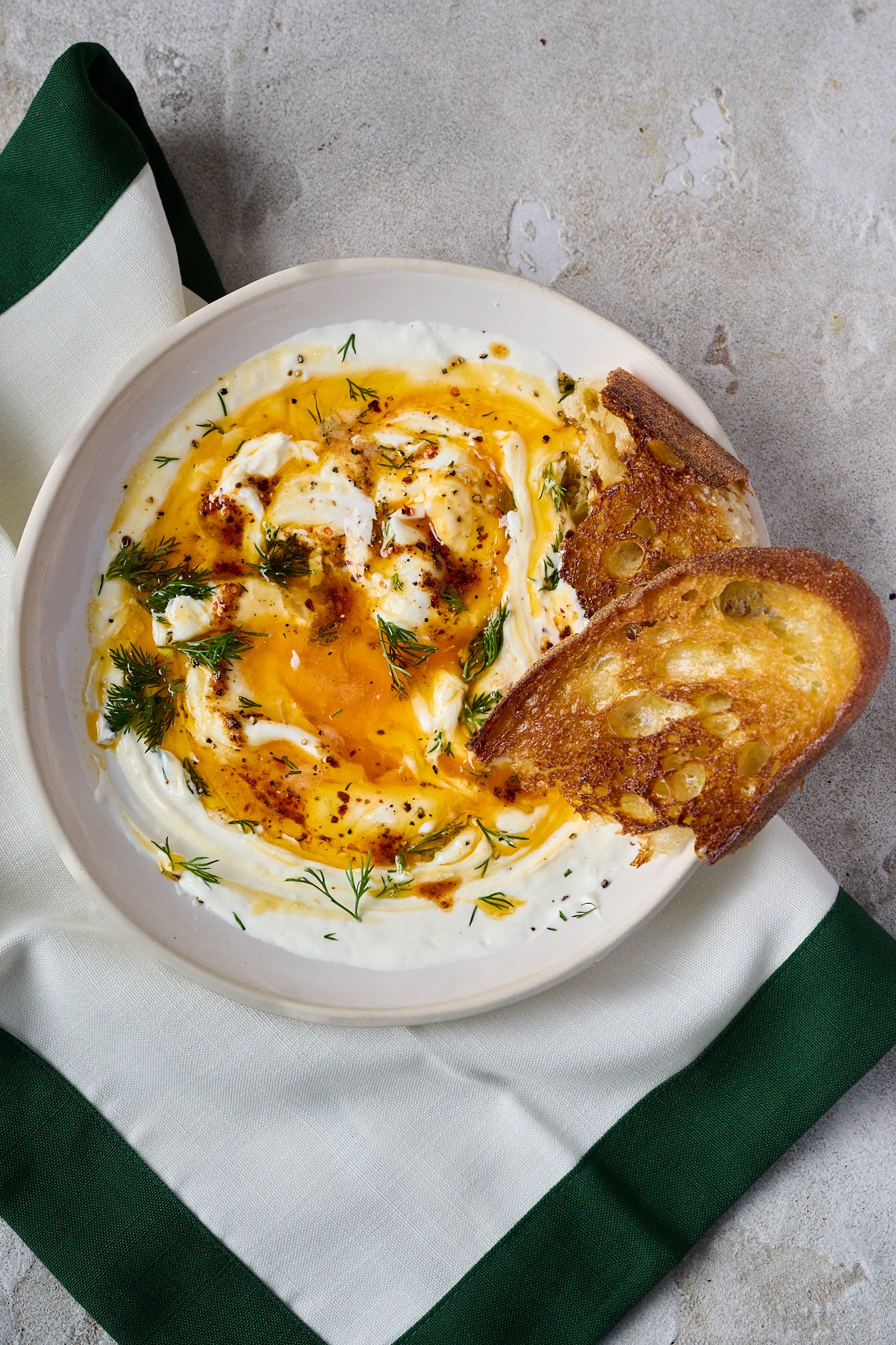 Turkish eggs are shown in white bowl. Two poached eggs have been broken open and sit on top of yoghurt with red butter sauce drizzled all over.  Two pieces of sourdough stick out the side of the bowl. 