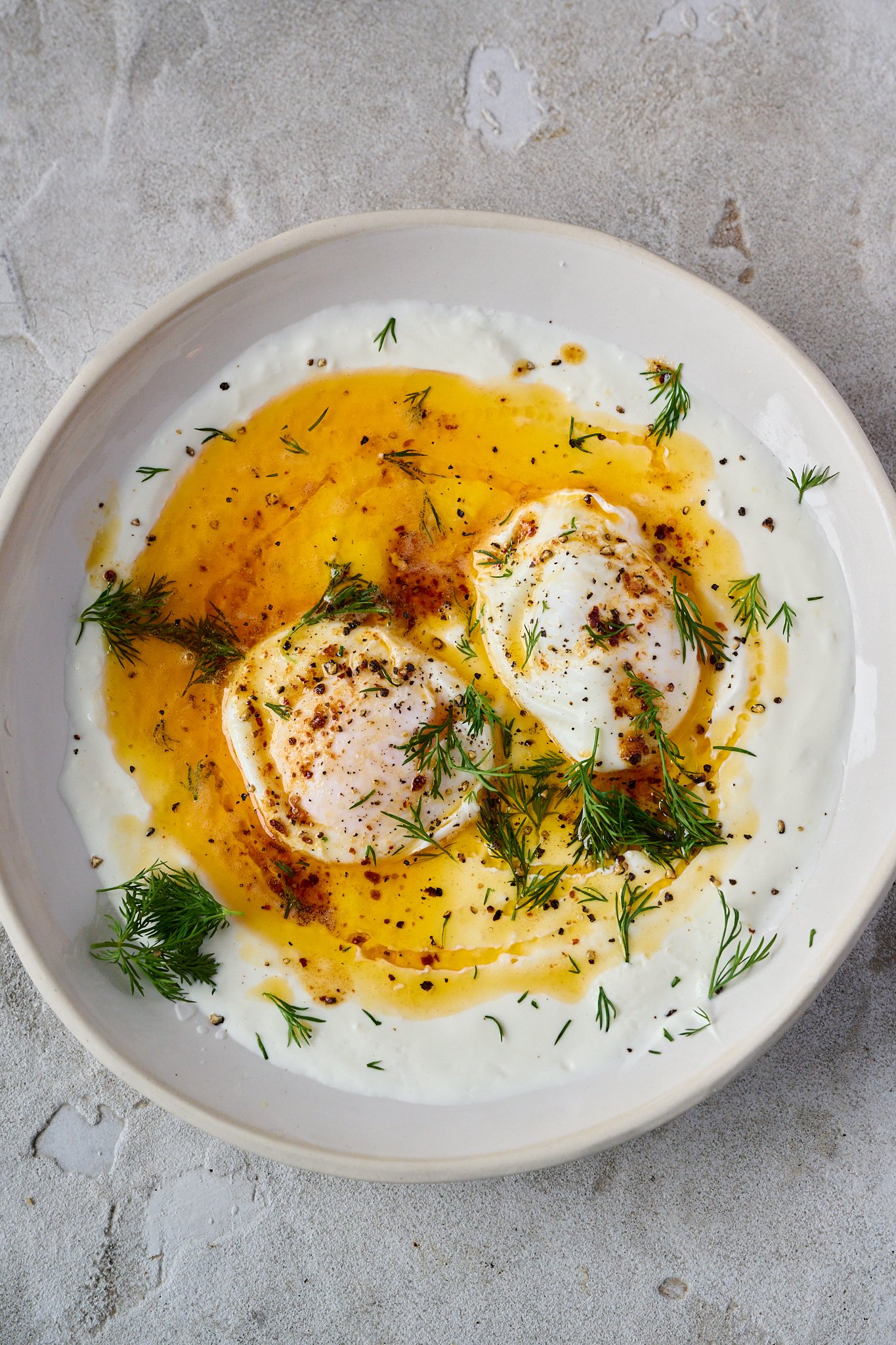 Turkish eggs are shown in white bowl. Two poached eggs sit on top of yoghurt with red butter sauce drizzled all over.