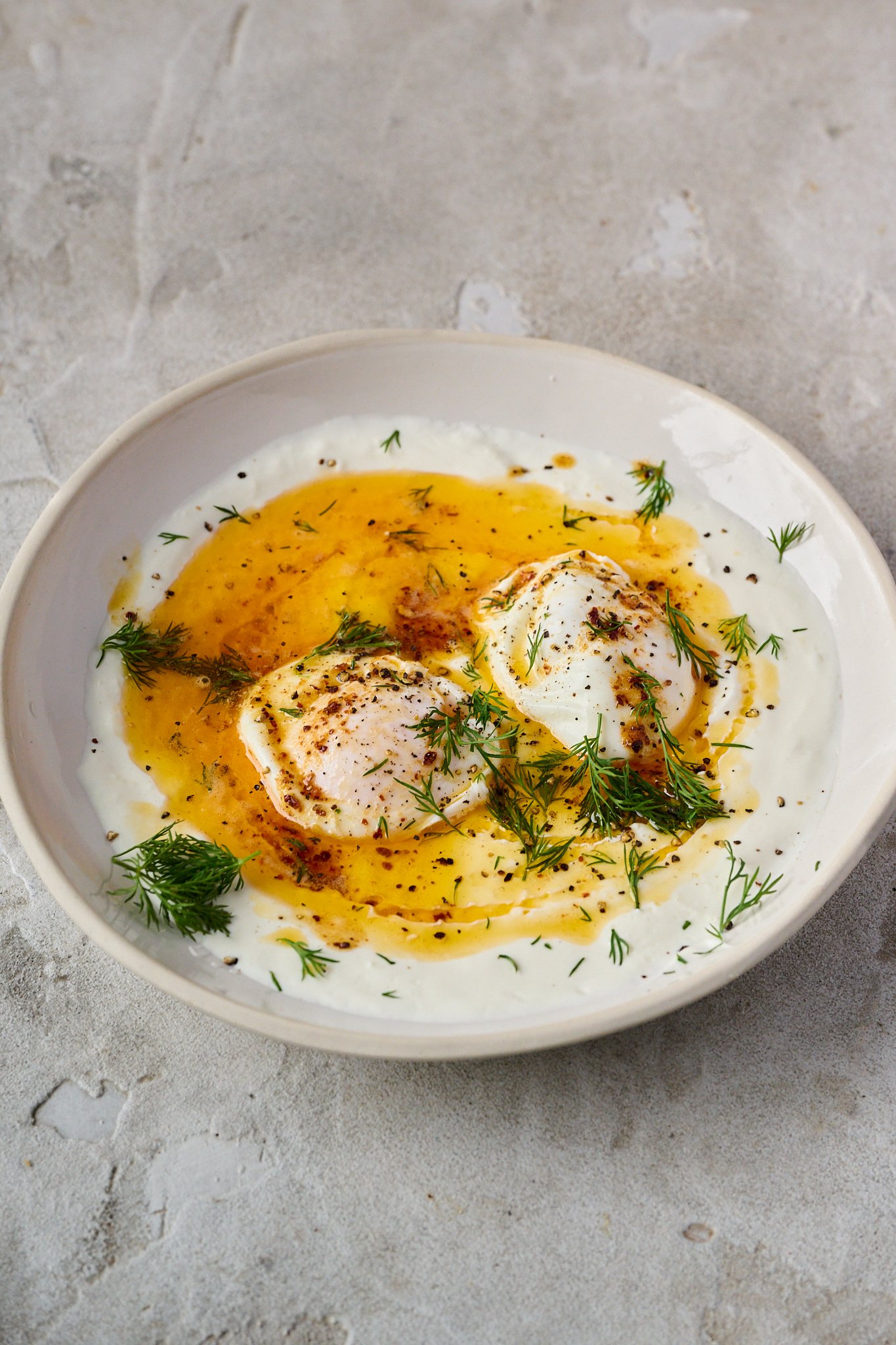 Turkish eggs are shown in white bowl. Two poached eggs sit on top of yoghurt with red butter sauce drizzled all over. 