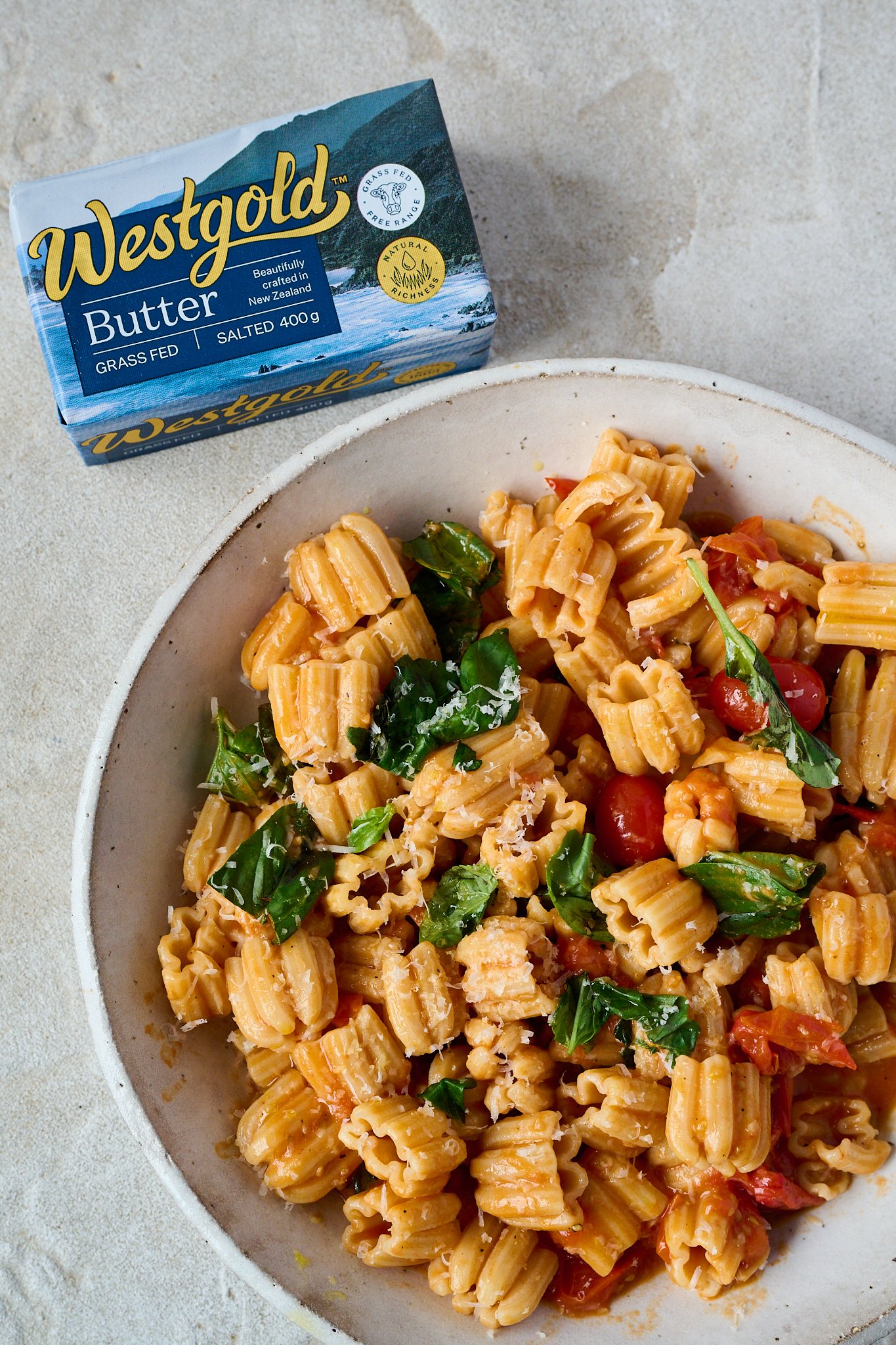 Tomato and butter pasta sits in large white bowl with fresh basil sprinkled all over and some grated parmesan cheese. You can see a slab of westgold butter in the top left hand corner. 