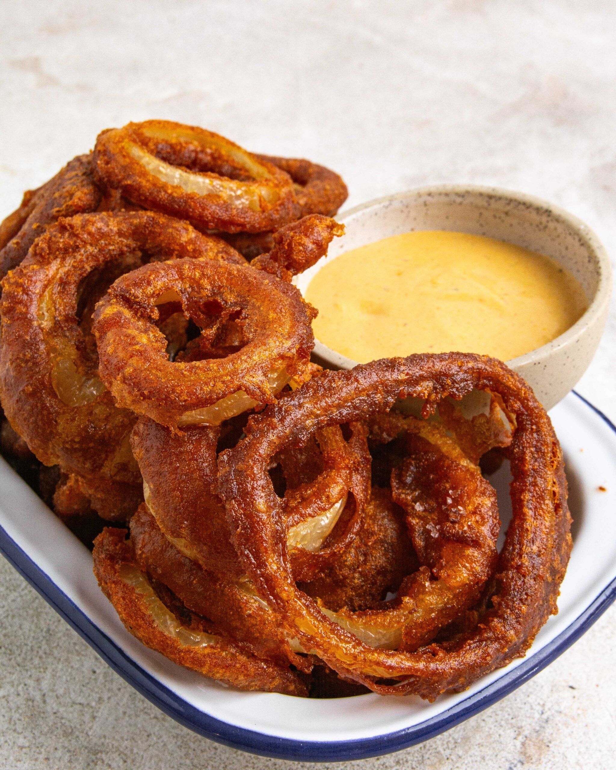 Beer battered onion rings sit in white tray with blue trim with a small bowl of dipping sauce to the side.