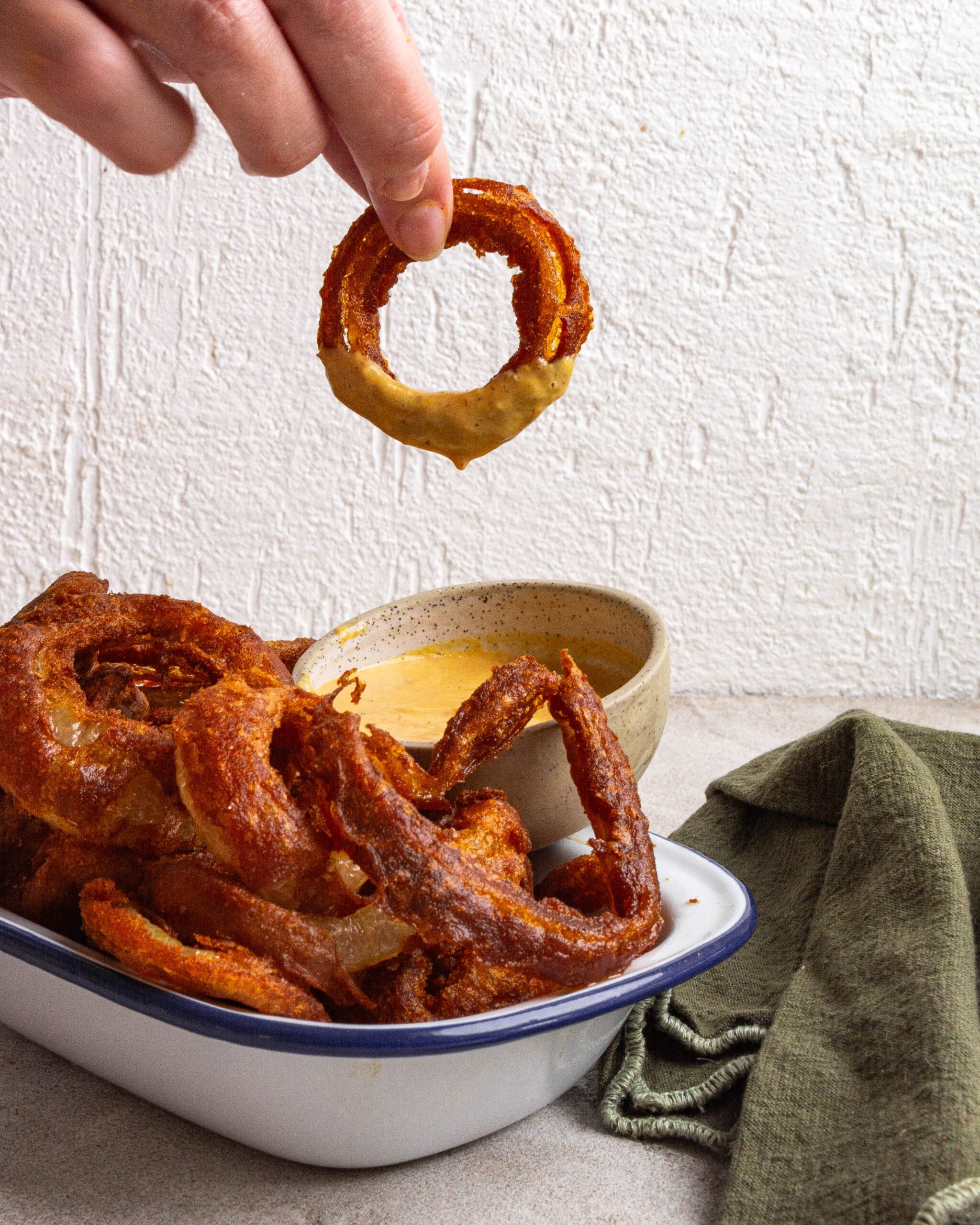 Beer battered onion rings sit in white tray with blue trim. One onion ring is being dipped into sauce. 