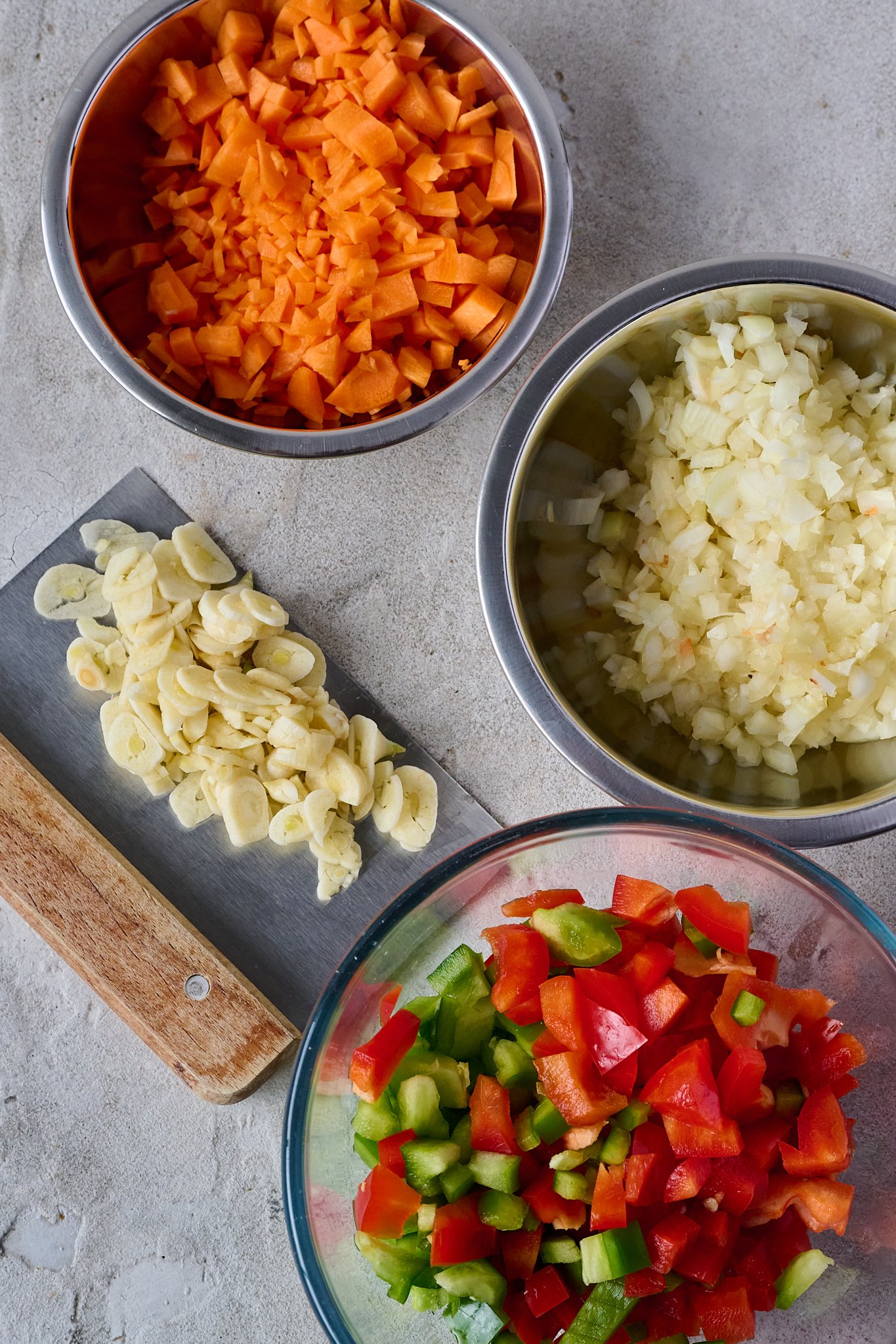 Chopped carrot, onion, capsicums and garlic sit in small stainless steel bowls. 