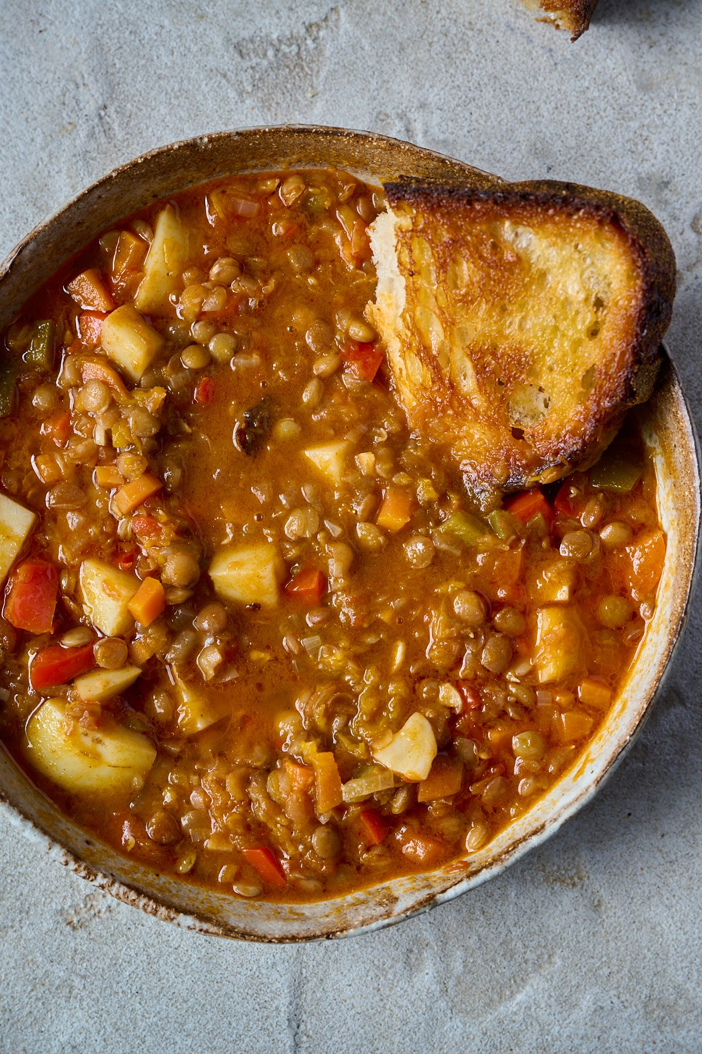 Spanish Lentil Stew (Lentejas) sits in brown bowl with piece of bread. 