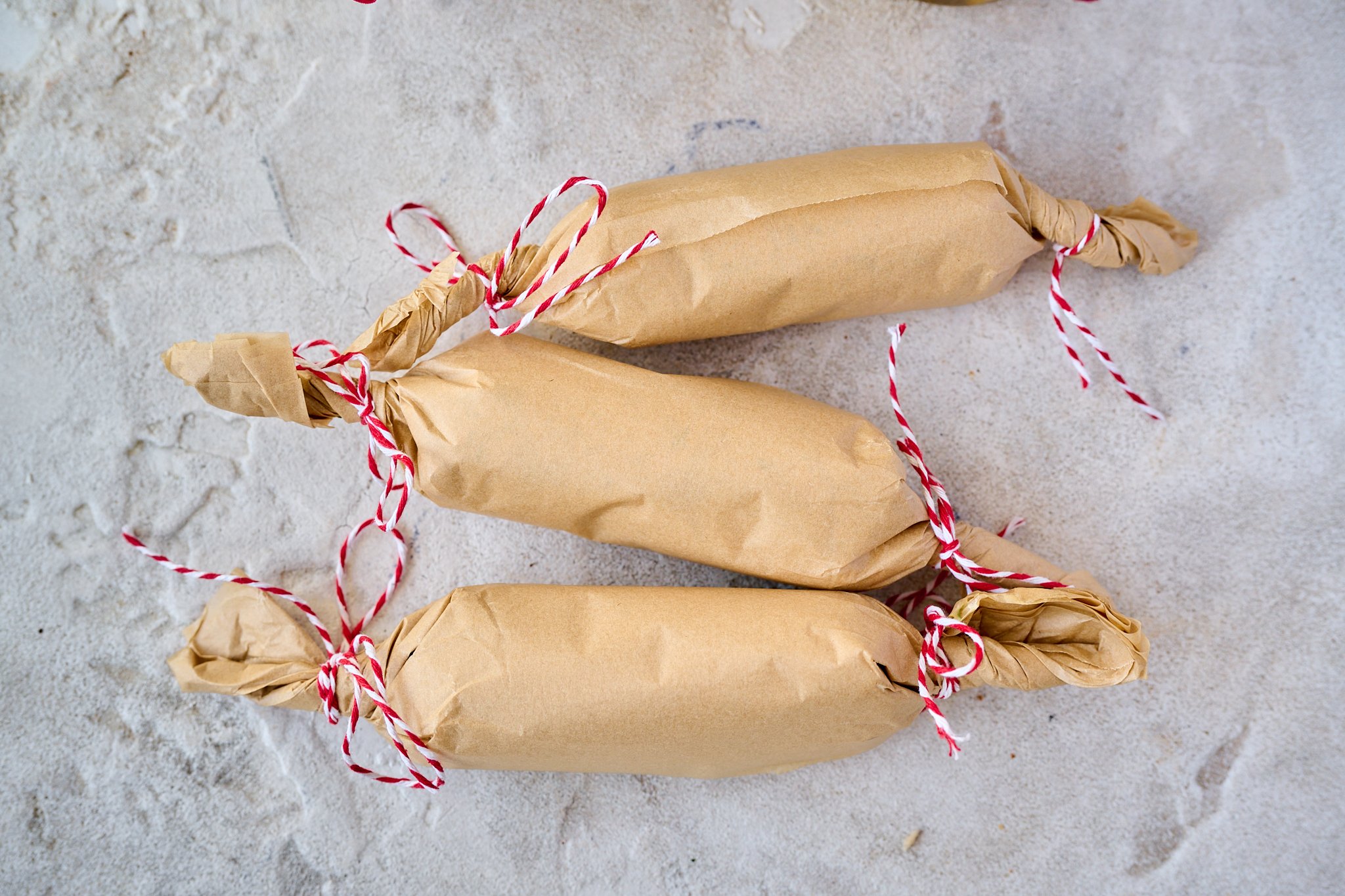 Homemade garlic butter is rolled into brown paper logs with red and white string tied at ends. 