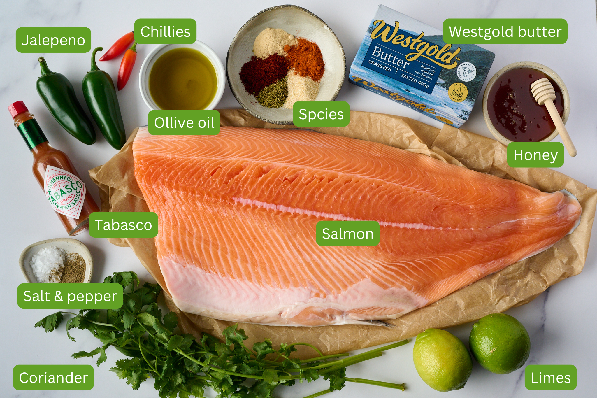 Ingredients for hot honey roasted salmon lay on marble counter. You can see large salmon, limes, salt and pepper, coriander, tabasco, olive oil. jalepeno, chillies, spices, honey and westgold butter