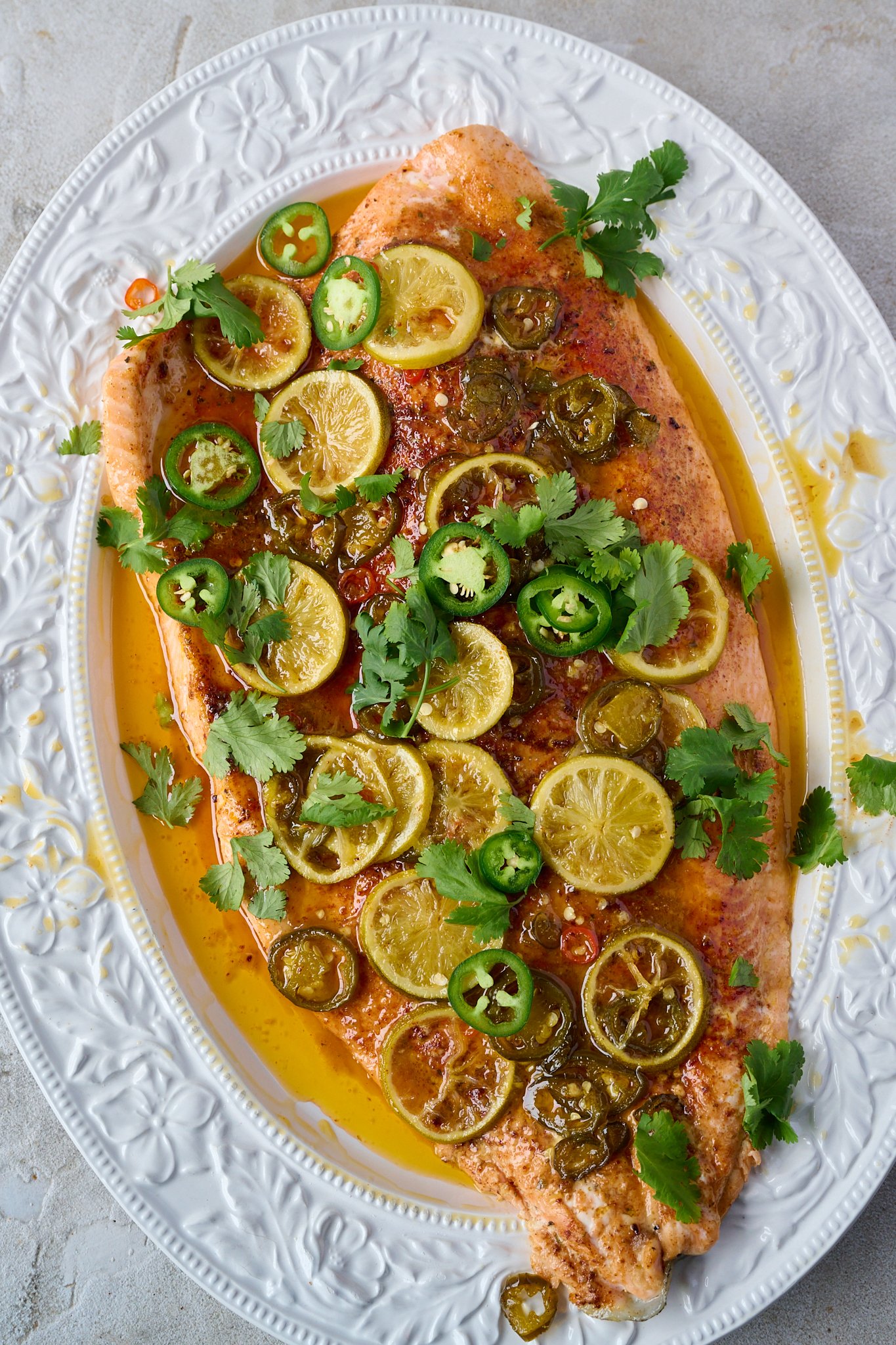 hot honey slow roasted salmon is in large white tray. Fresh coriander is sprinkled all over and you can see the cooked limes and jalepeno.