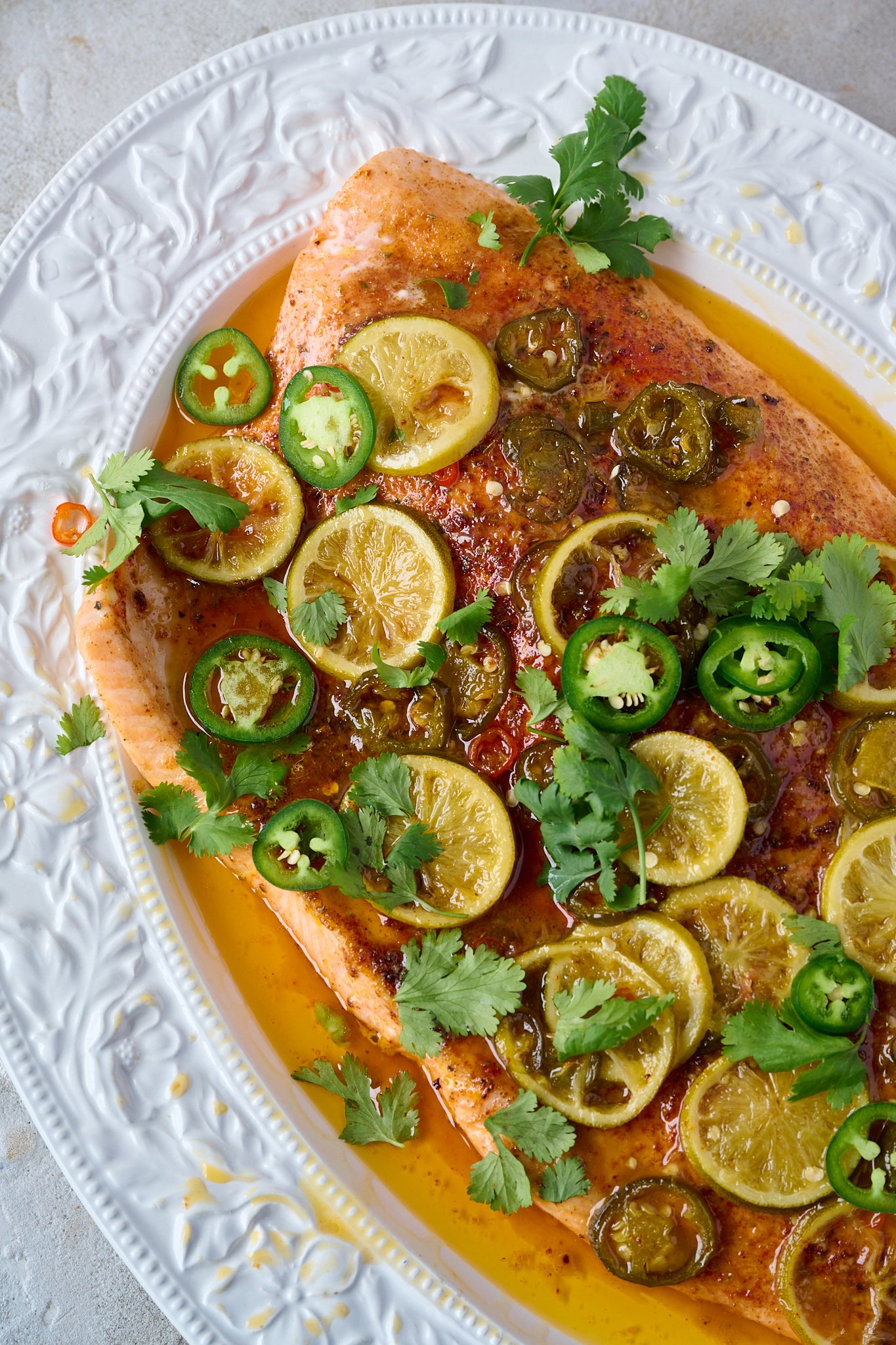hot honey slow roasted salmon is in large white tray. Fresh coriander is sprinkled all over and you can see the cooked limes and jalepeno.