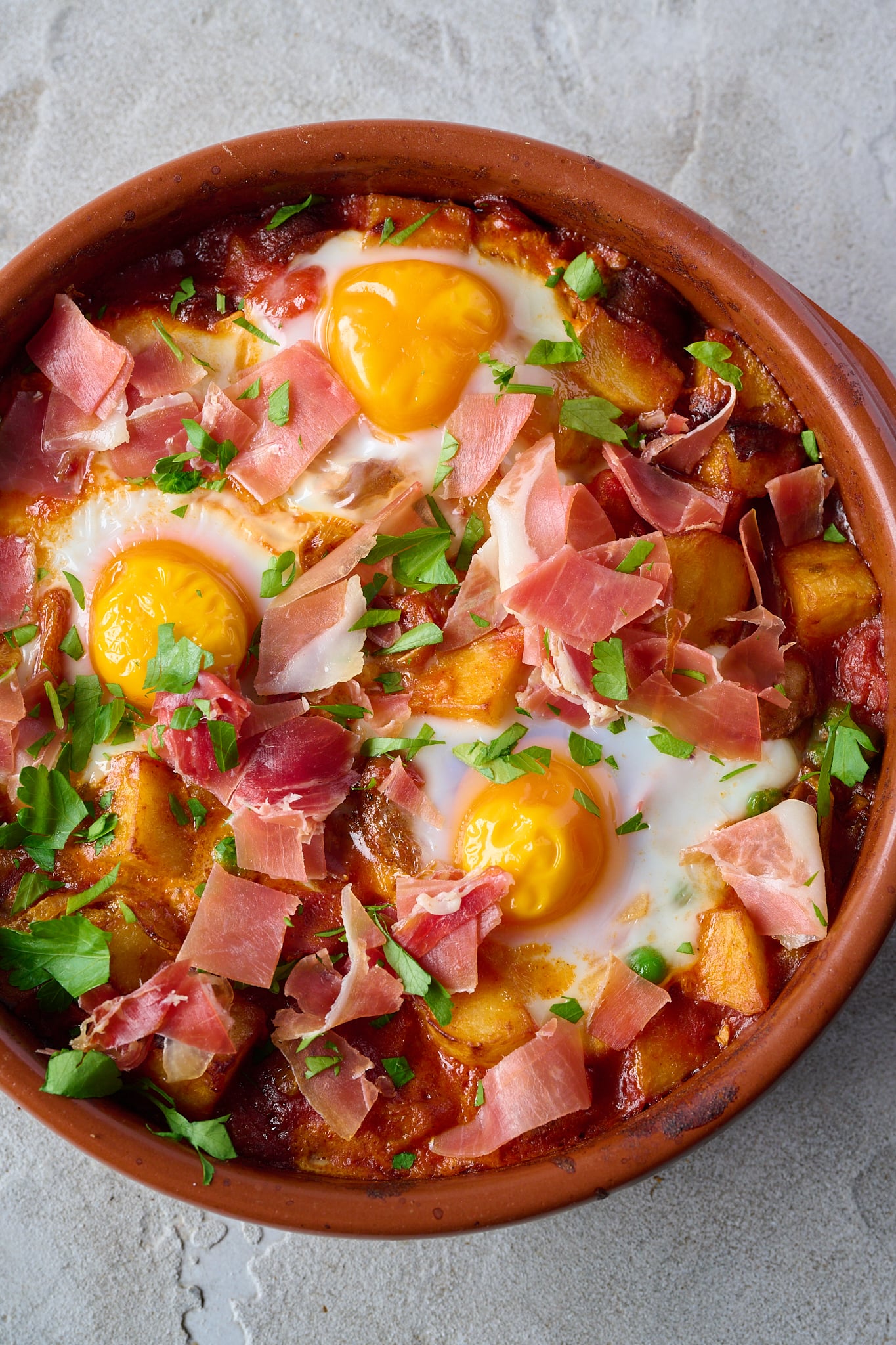 Spanish Baked Eggs (Huevos a la Flamenca) are in clay pot with jamon and parsley sprinkled over.