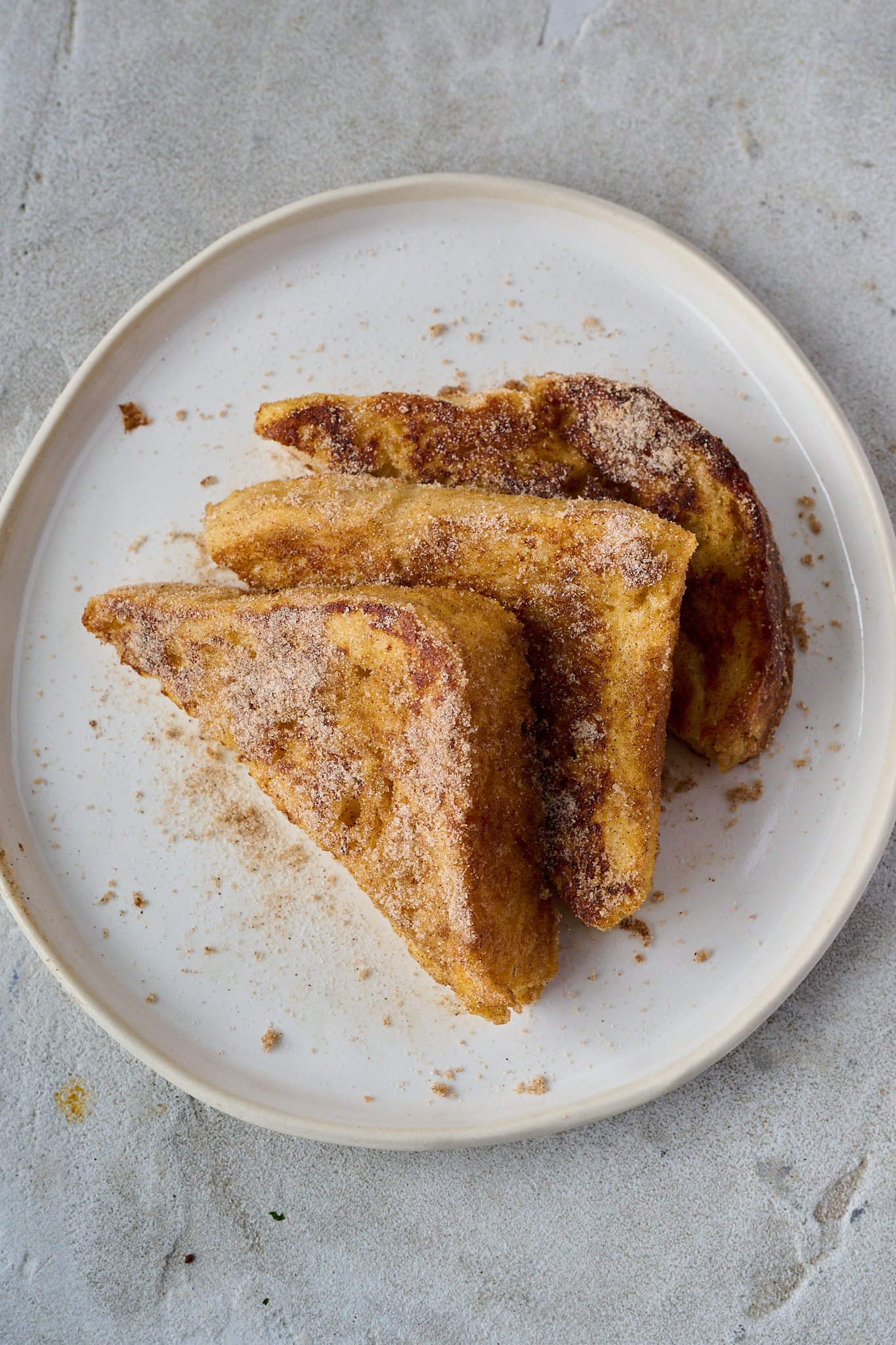 Three slices of Torrijas (Spanish French Toast) sit on a white plate. They are dusted in lots of cinnamon sugar. 