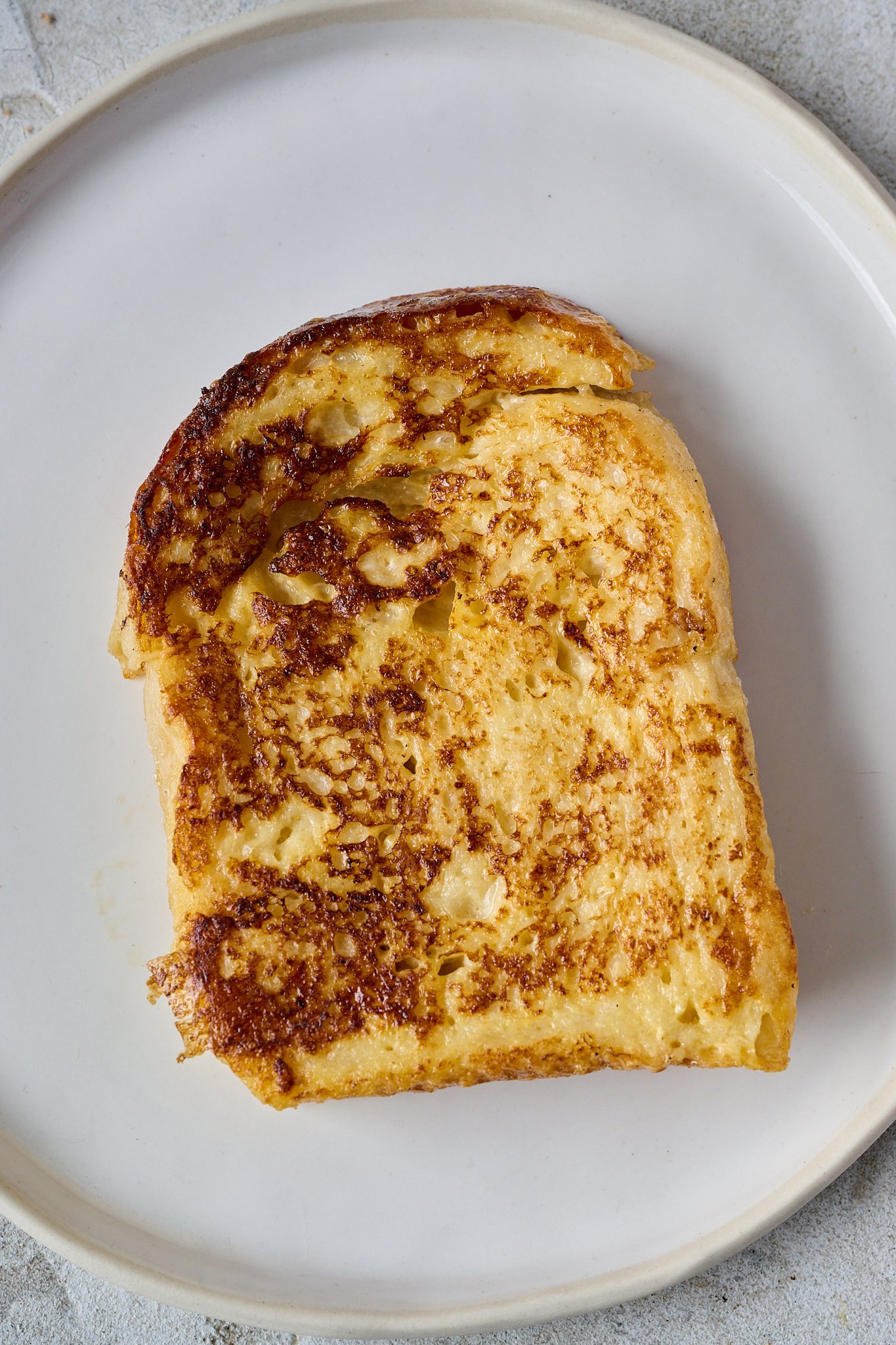 A thick slice of white bread has been fried and is sitting on a white plate. It is crispy and golden on the outside.