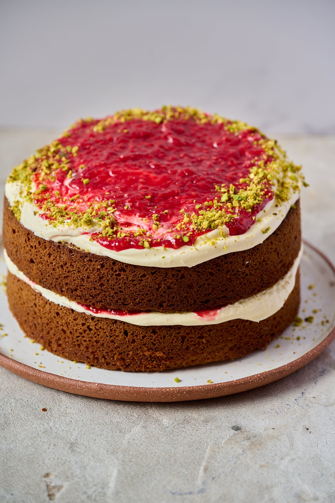 White Chocolate, Pistachio & Raspberry Olive Oil Cake sits on a white plate with a brown rim.