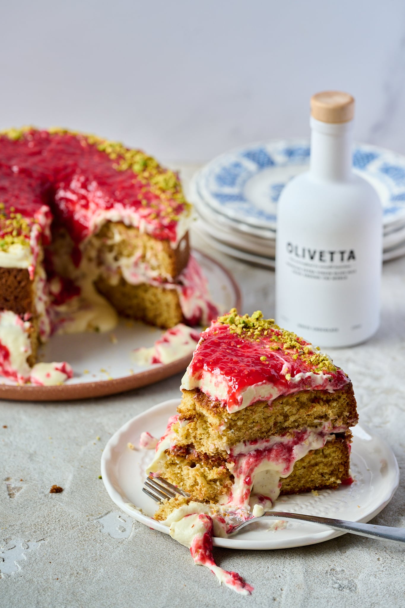 A slice of White Chocolate, Pistachio & Raspberry Olive Oil Cake sits on a white plate with a fork. You can see the large cake in the background and the white chocolate bottle of olive oil