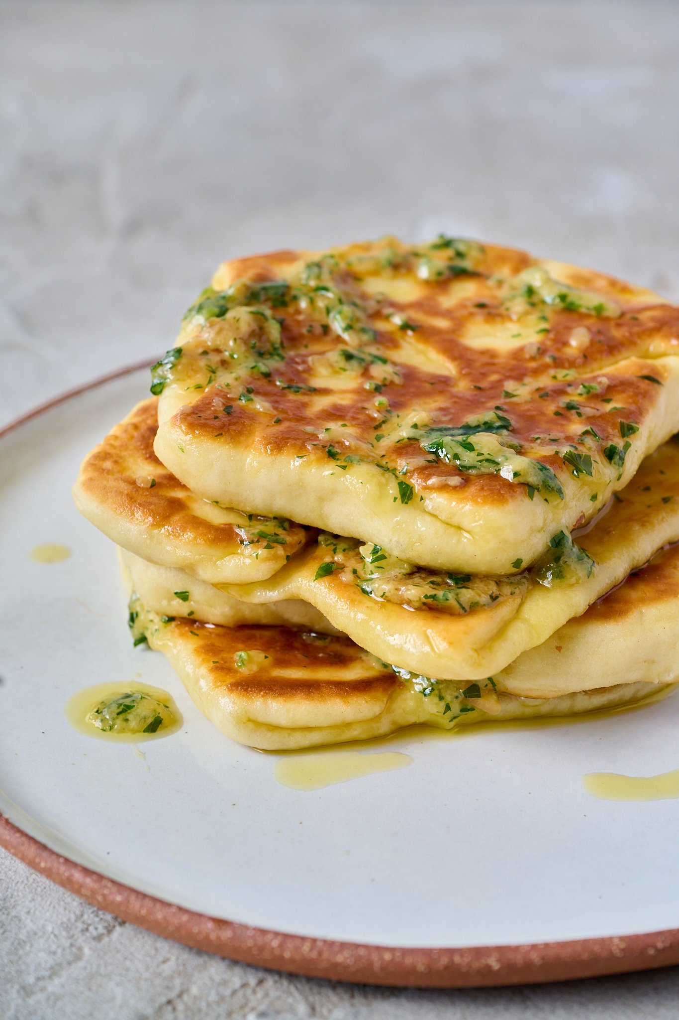 3 Ingredient yogurt flatbreads are piled on top of each other and sit on a white plate. They are drizzled with garlic butter.