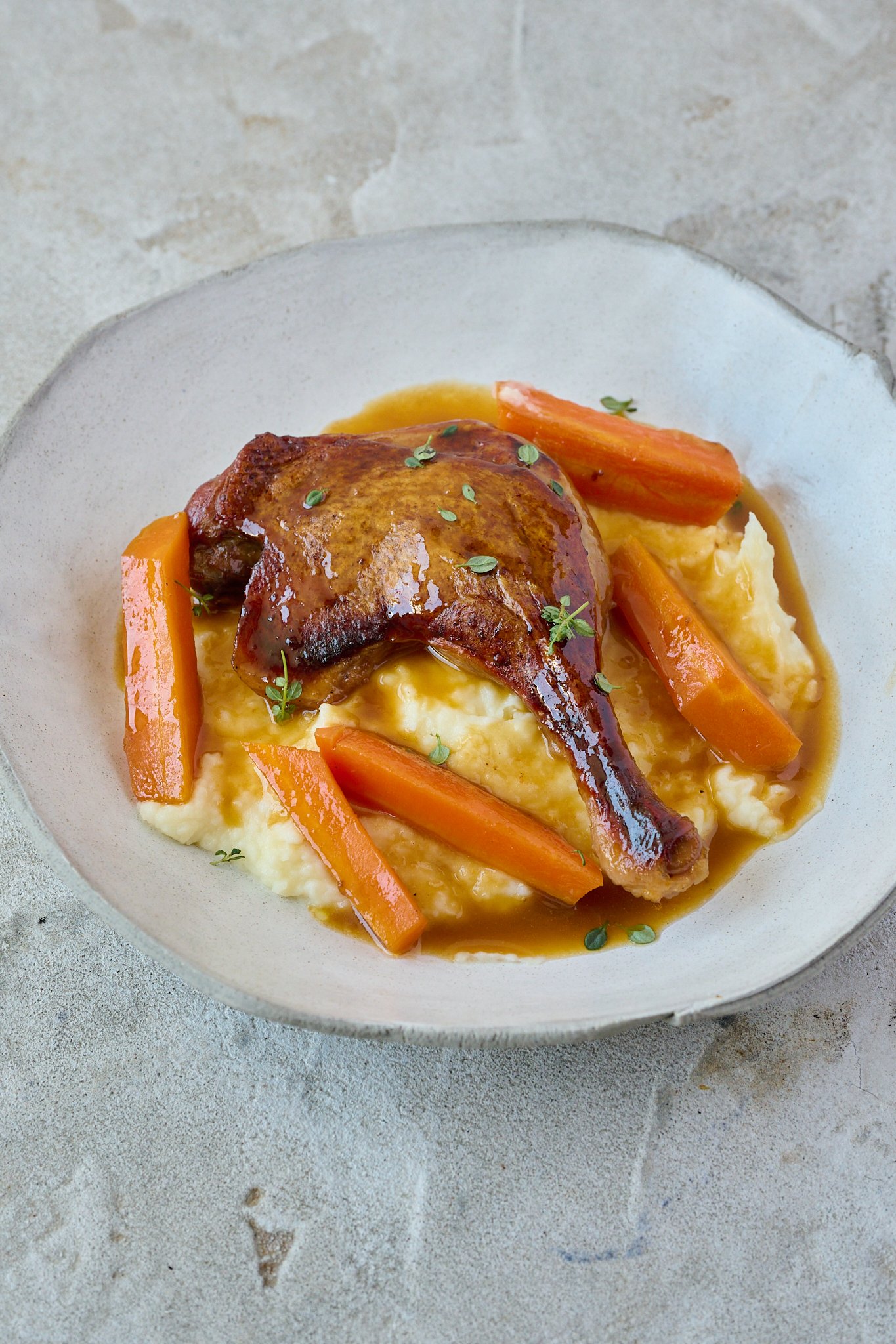Crispy Braised Duck Legs with Spiced Orange Sauce sits on pile of mash potatoes in large white bowl. Fresh thyme leaves is sprinkled all over.