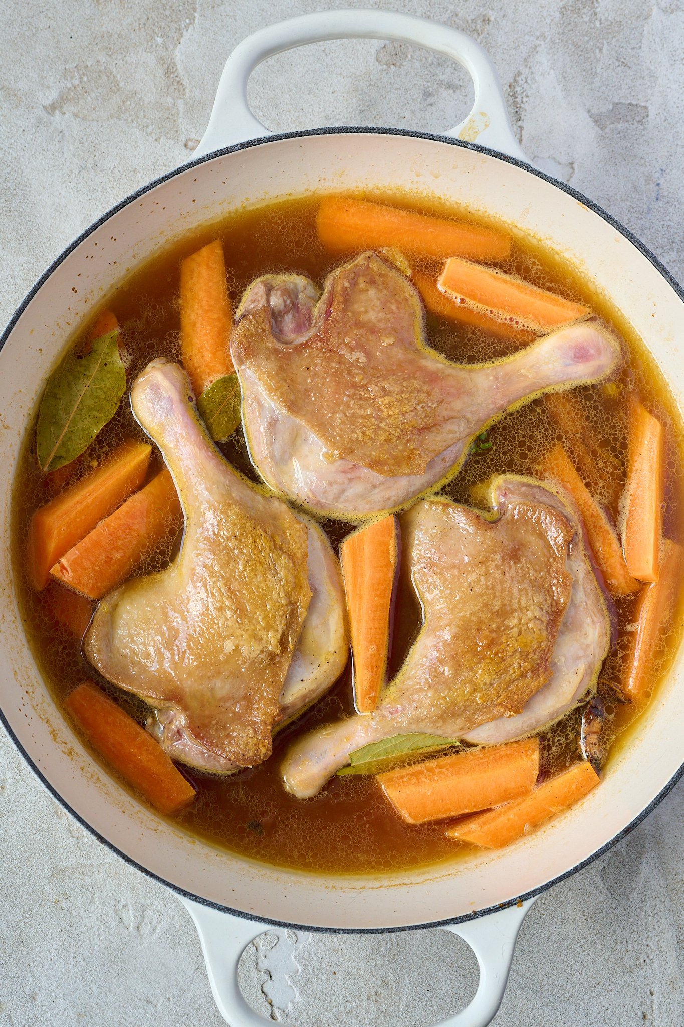 Three duck legs sits in a white pan with a brown sauce and carrots.