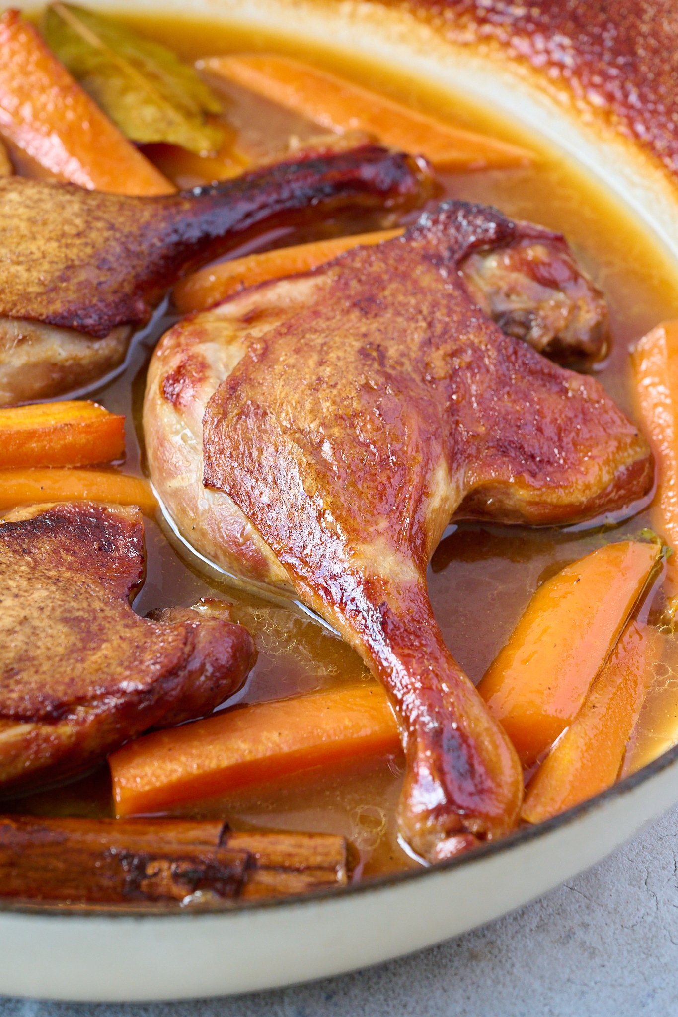 Close up image of crispy duck leg is shown in a white pan with brown sauce and carrots.