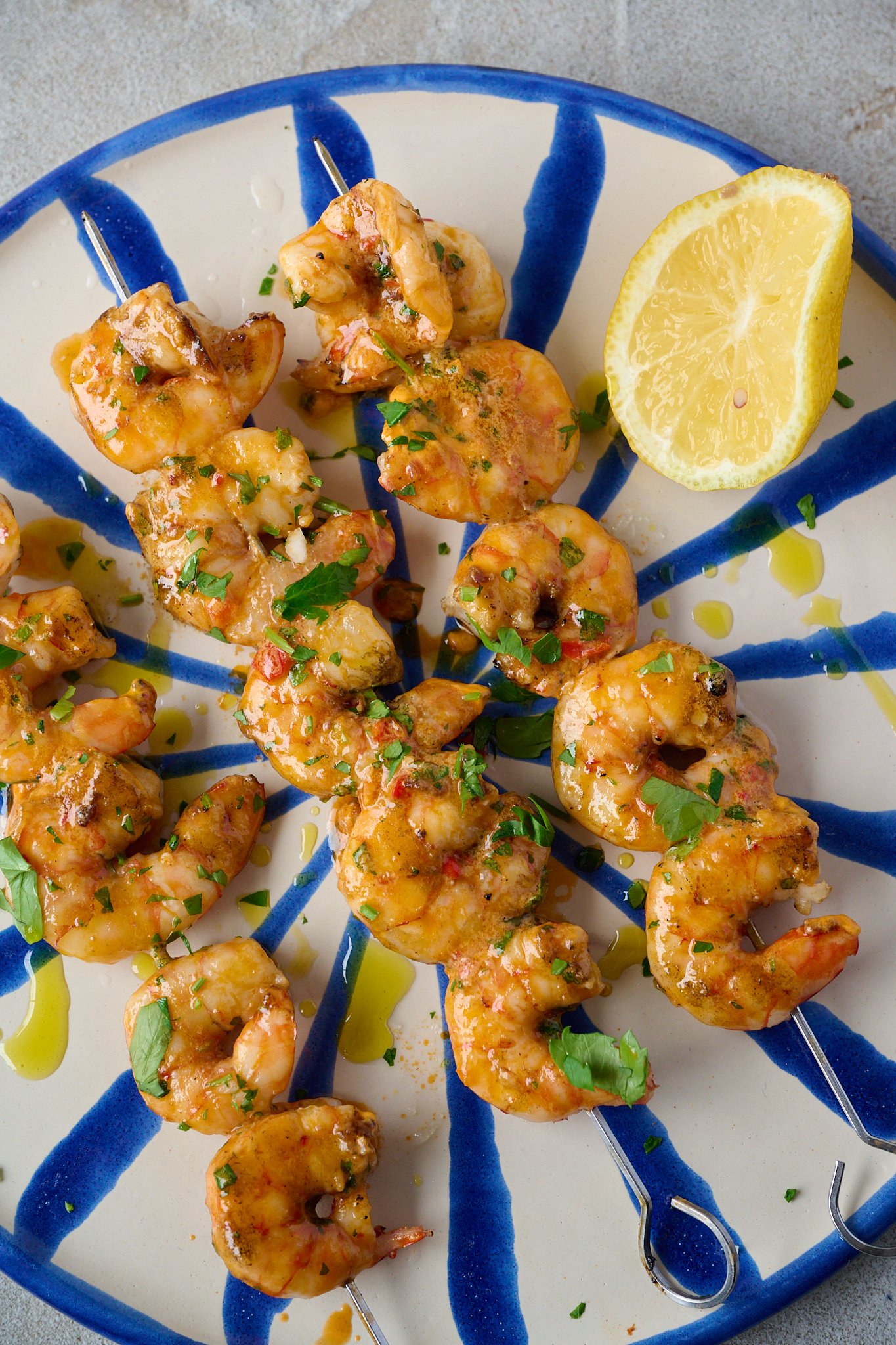 Grilled Shrimp Skewers with Tomato Butter (BBQ Prawns) are on blue and white striped plate with fresh parsley sprinkled over. A lemon sits on the right side of the plate.