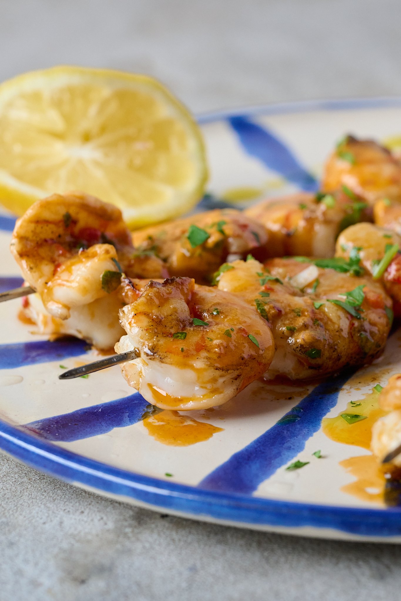 Grilled Shrimp Skewers with Tomato Butter (BBQ Prawns) are on blue and white striped plate with fresh parsley sprinkled over. A lemon sits on the in the left side of the plate.