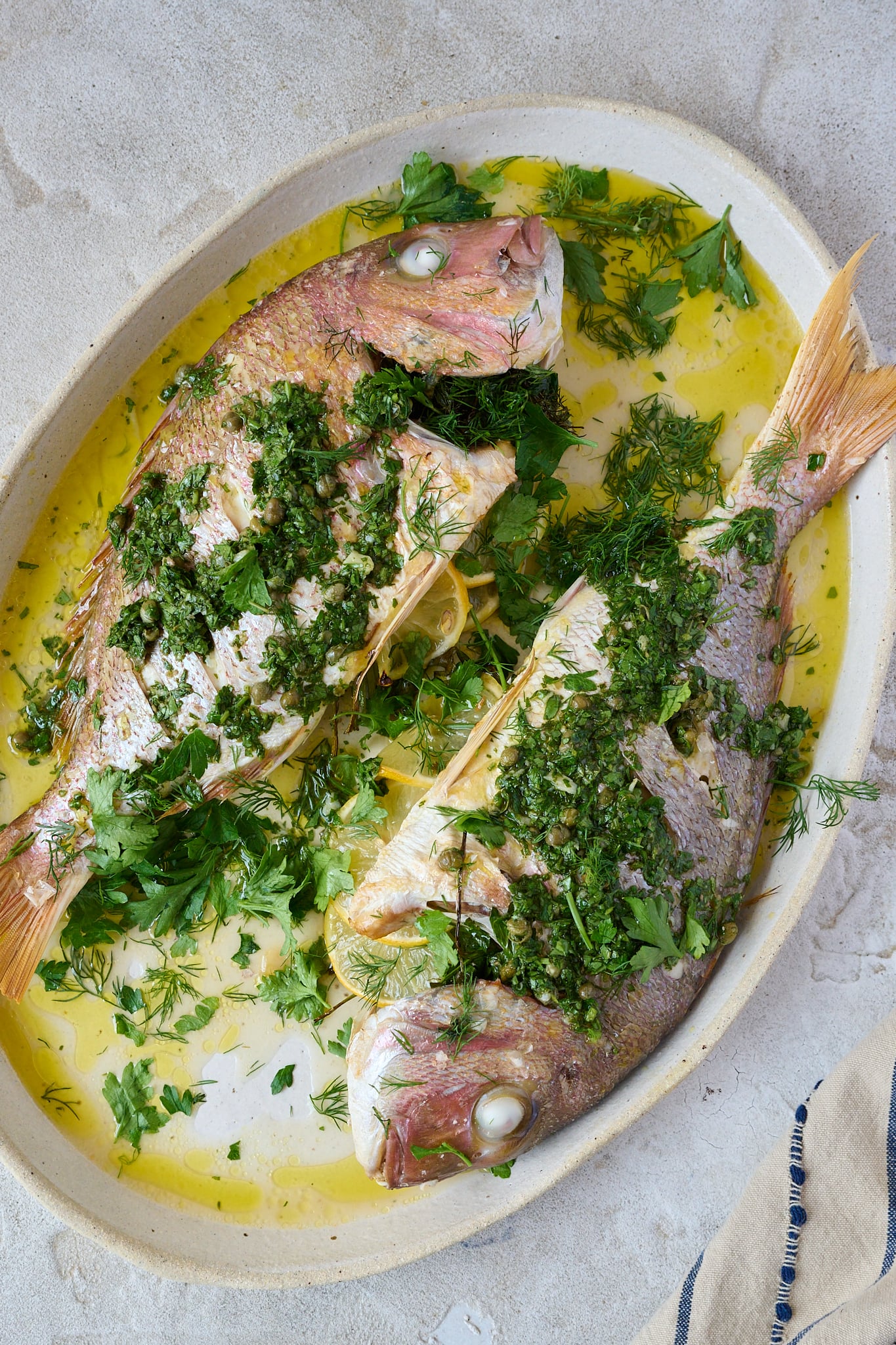 Whole baked fish with salsa verde sits on large white platter. Lots of herbs are scattered all over. 