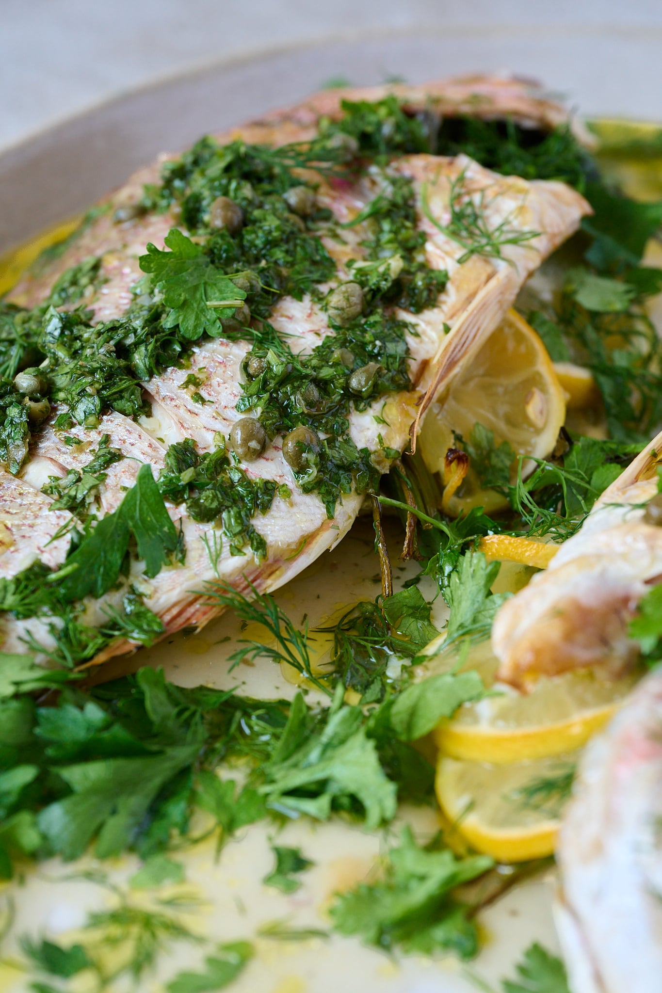 Whole baked fish with salsa verde sits on large white platter. Lots of herbs are scattered all over.