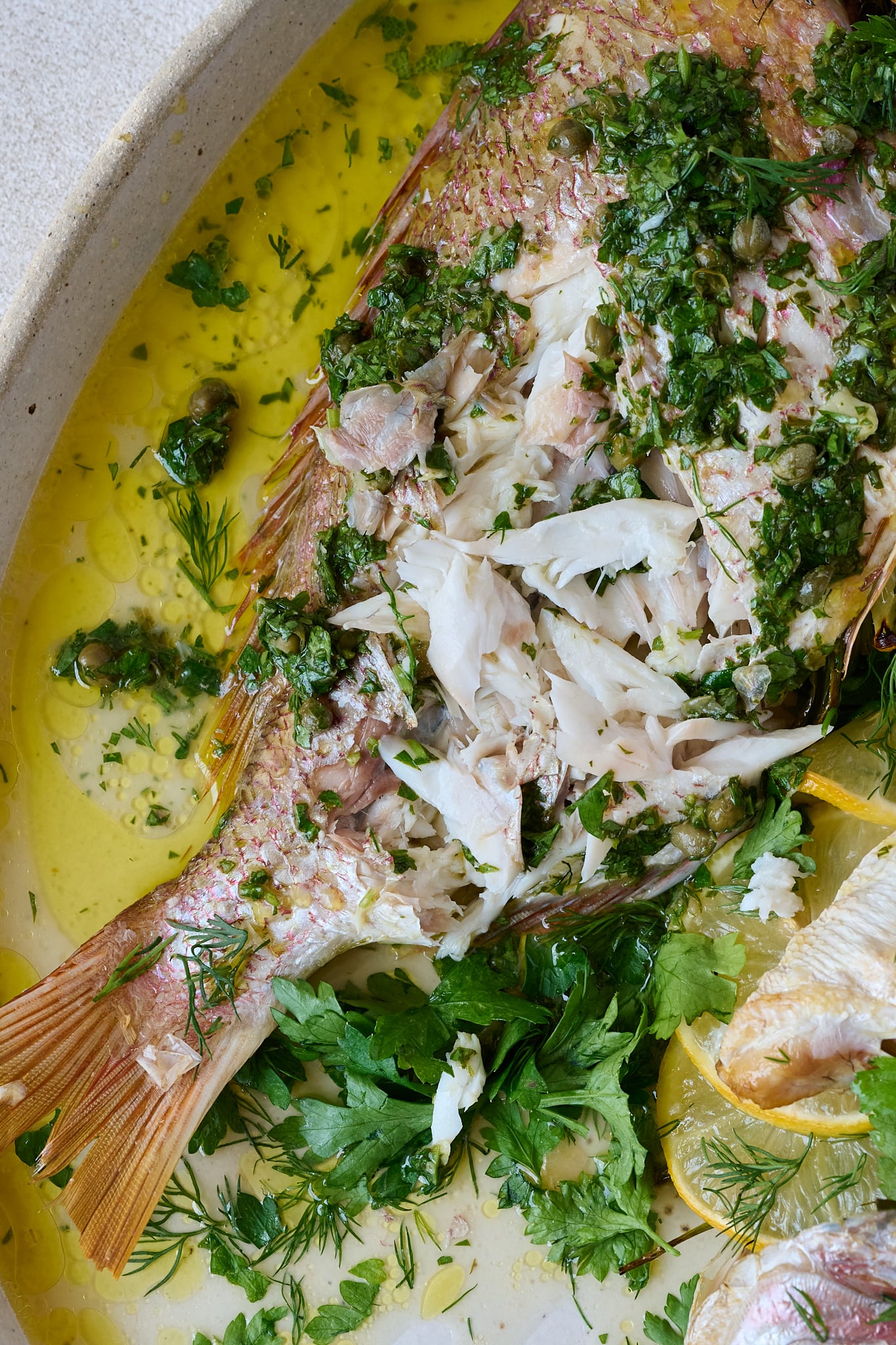 Whole baked fish with salsa verde sits on large white platter. Lots of herbs are scattered all over. you can see flesh of fish has been broken into.