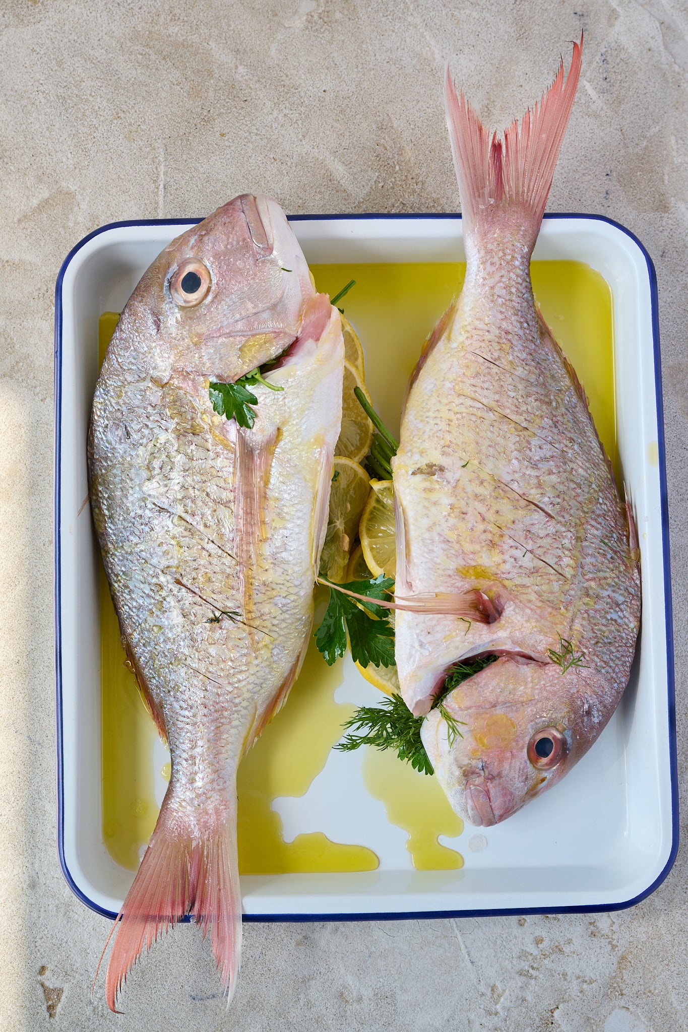 Whole baked snapper is sitting on white baking tray. Has herbs poking out of belly and olive oil drizzled all over.