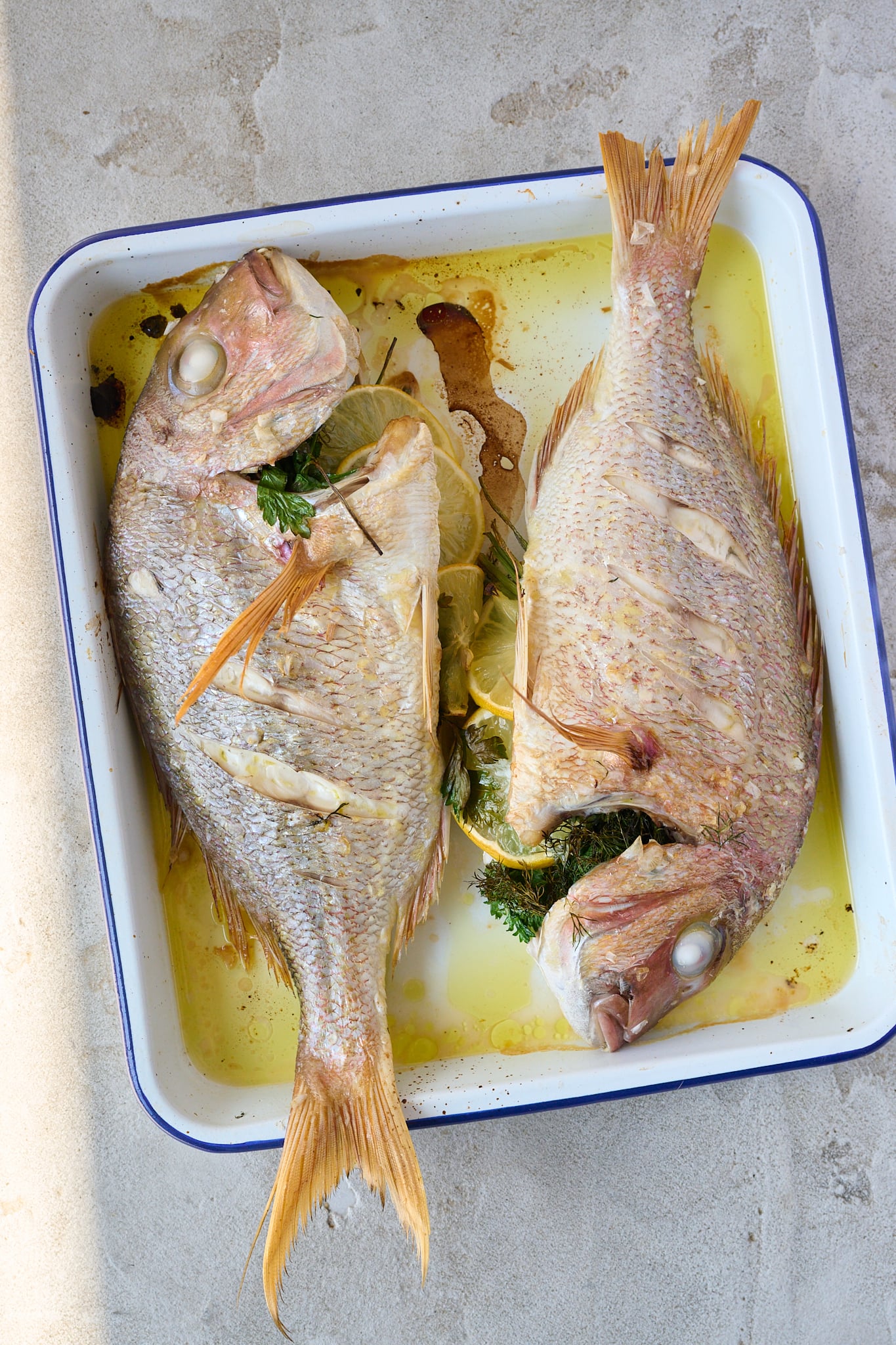Whole baked fish with salsa verde has been roasted and sits on large white platter. Lots of herbs are scattered all over.